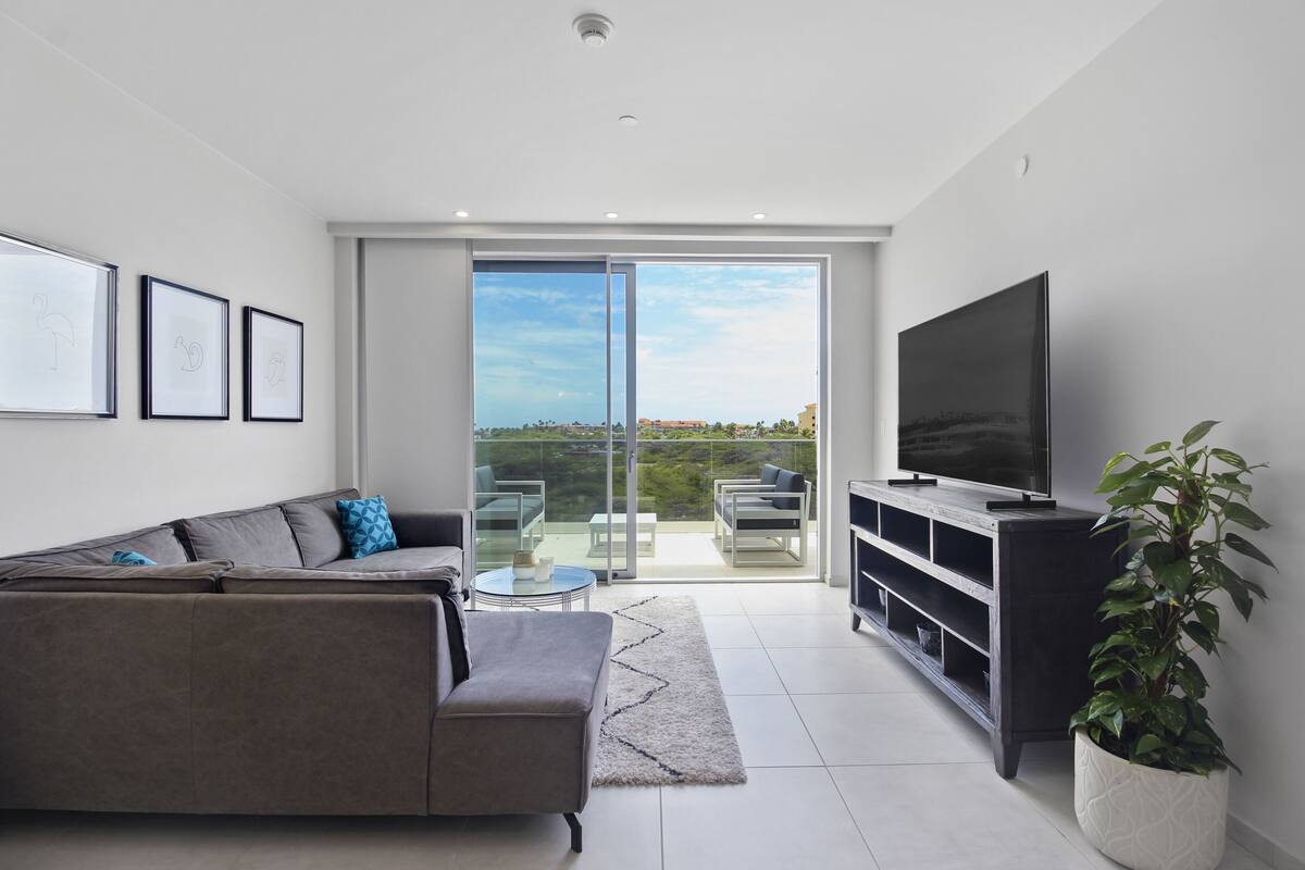 A modern living room with a grey sofa, a flat-screen TV, a plant, and a sliding glass door leading to a balcony.