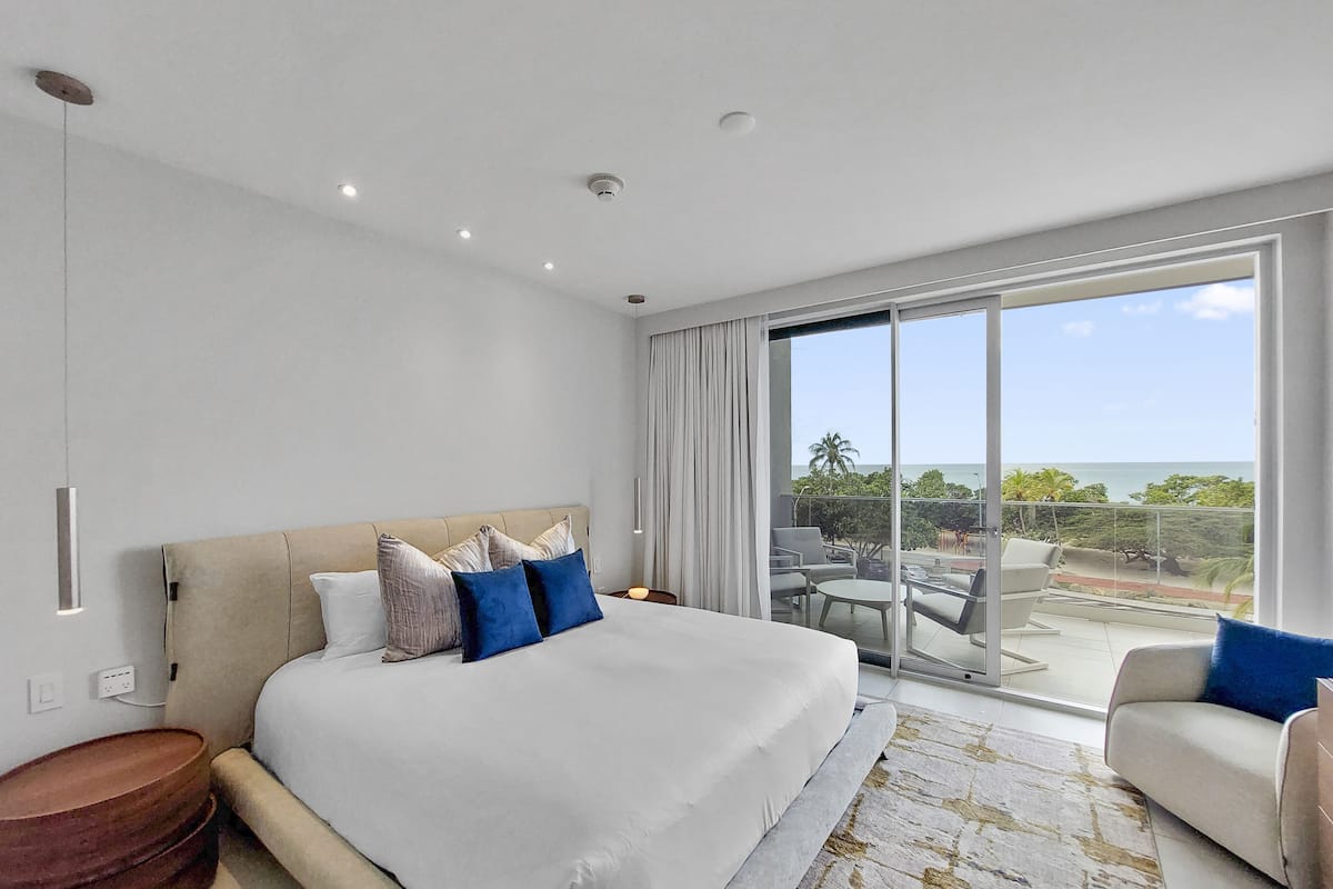 A modern hotel room with a large bed, a balcony with outdoor seating, and a view of the ocean.