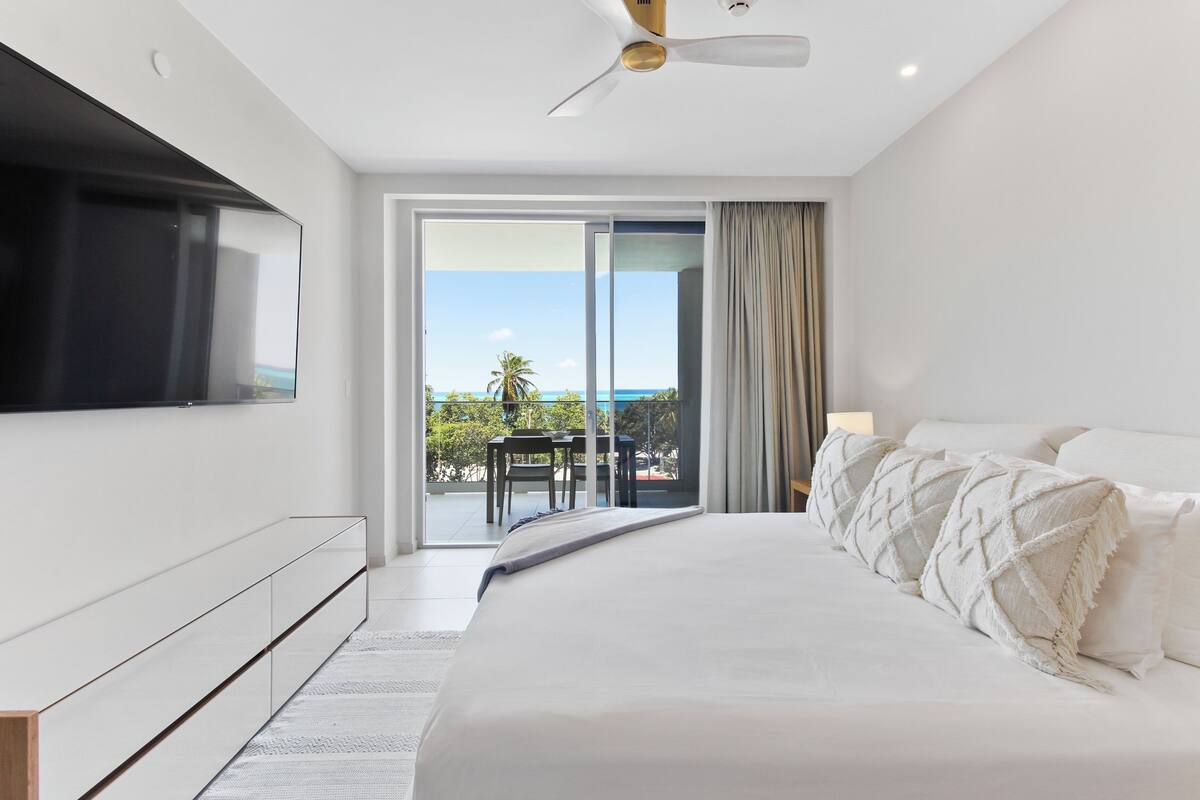 A modern hotel room with a large bed, a flat-screen TV mounted on the wall, and a balcony with a view of the ocean.