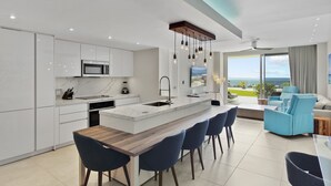 Una cucina moderna con mobili bianchi, un'isola centrale con sgabelli da bar e vista sull'oceano attraverso ampie finestre.