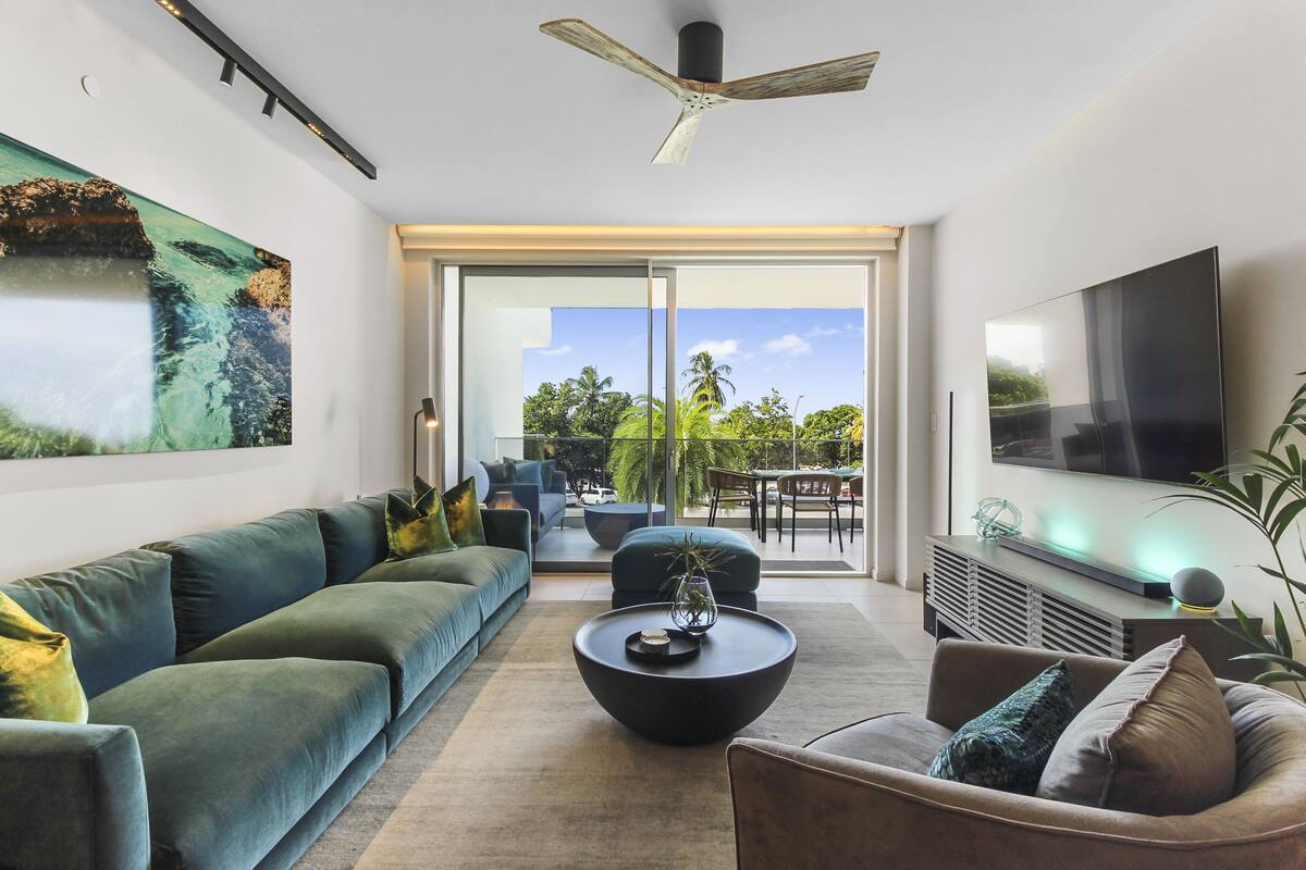 A modern living room with a large sofa, a coffee table, and a flat-screen TV. There is a sliding glass door leading to an outdoor area with a view of trees and a clear sky.