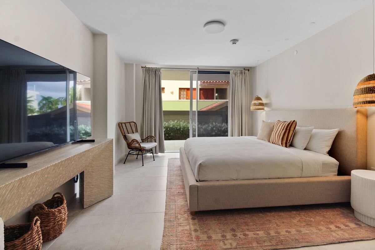 A modern bedroom with a large bed, a TV on a wooden console, a wicker chair, and a view through a sliding door.