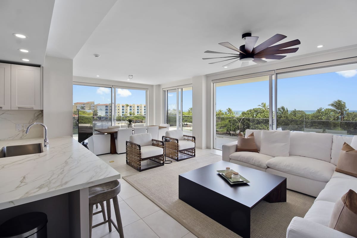 A modern living room with a white sofa, a coffee table, and a kitchen area with a marble countertop and white cabinets.