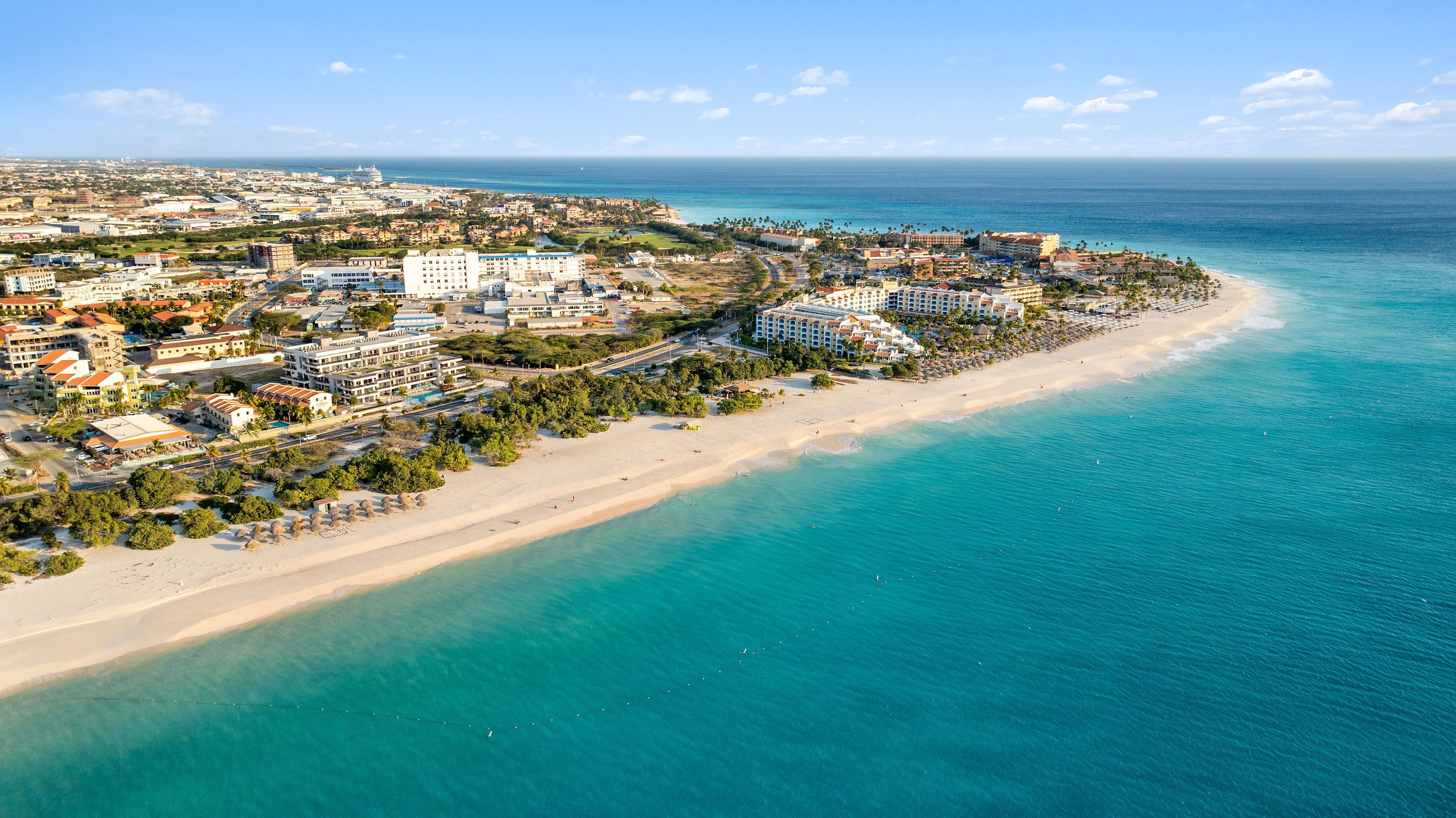 A coastal cityscape with a beachfront hotel, clear blue waters, and a mix of residential and commercial buildings.