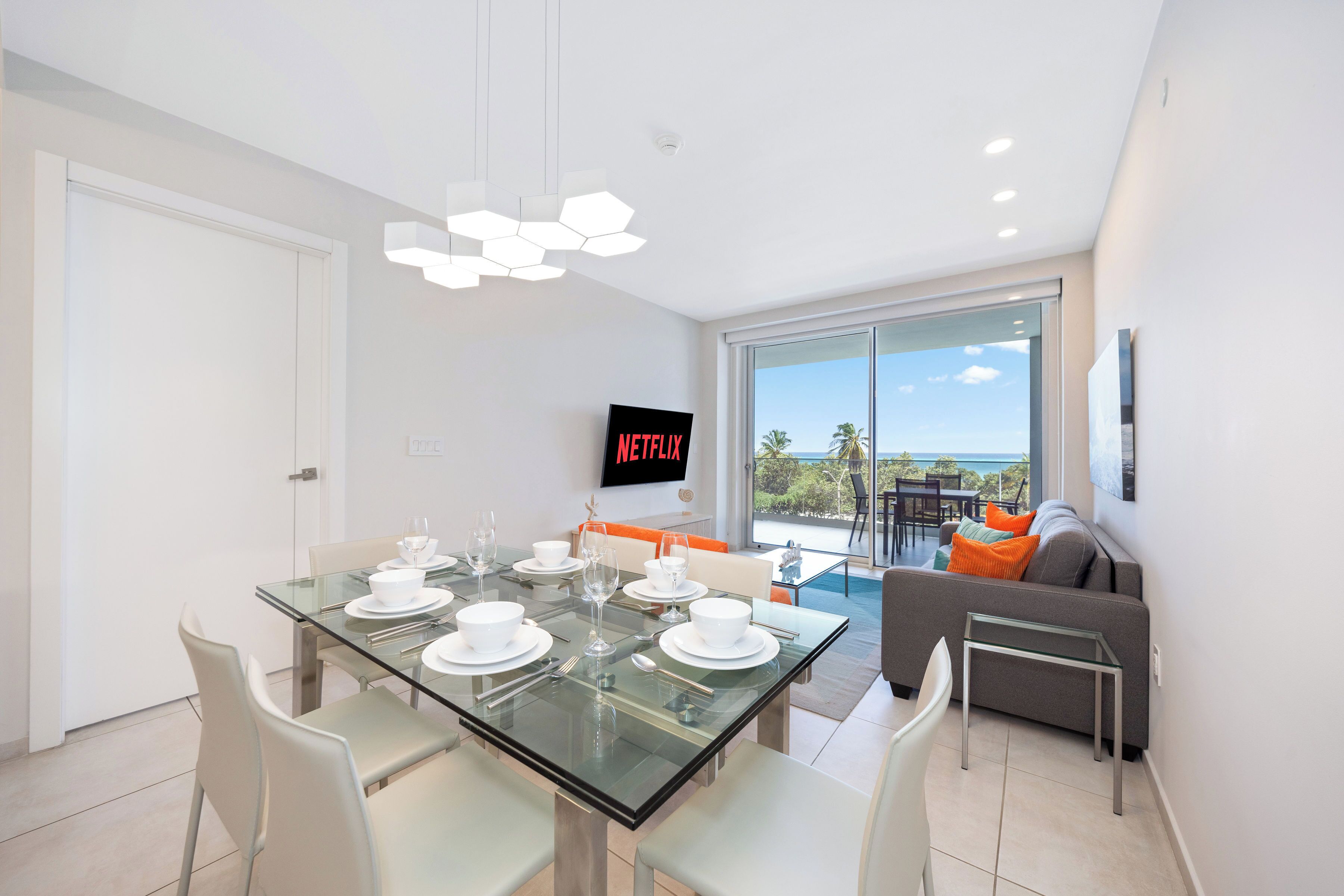 A modern dining area with a glass table set for six, a sofa with orange cushions, and a view of the ocean through large windows.