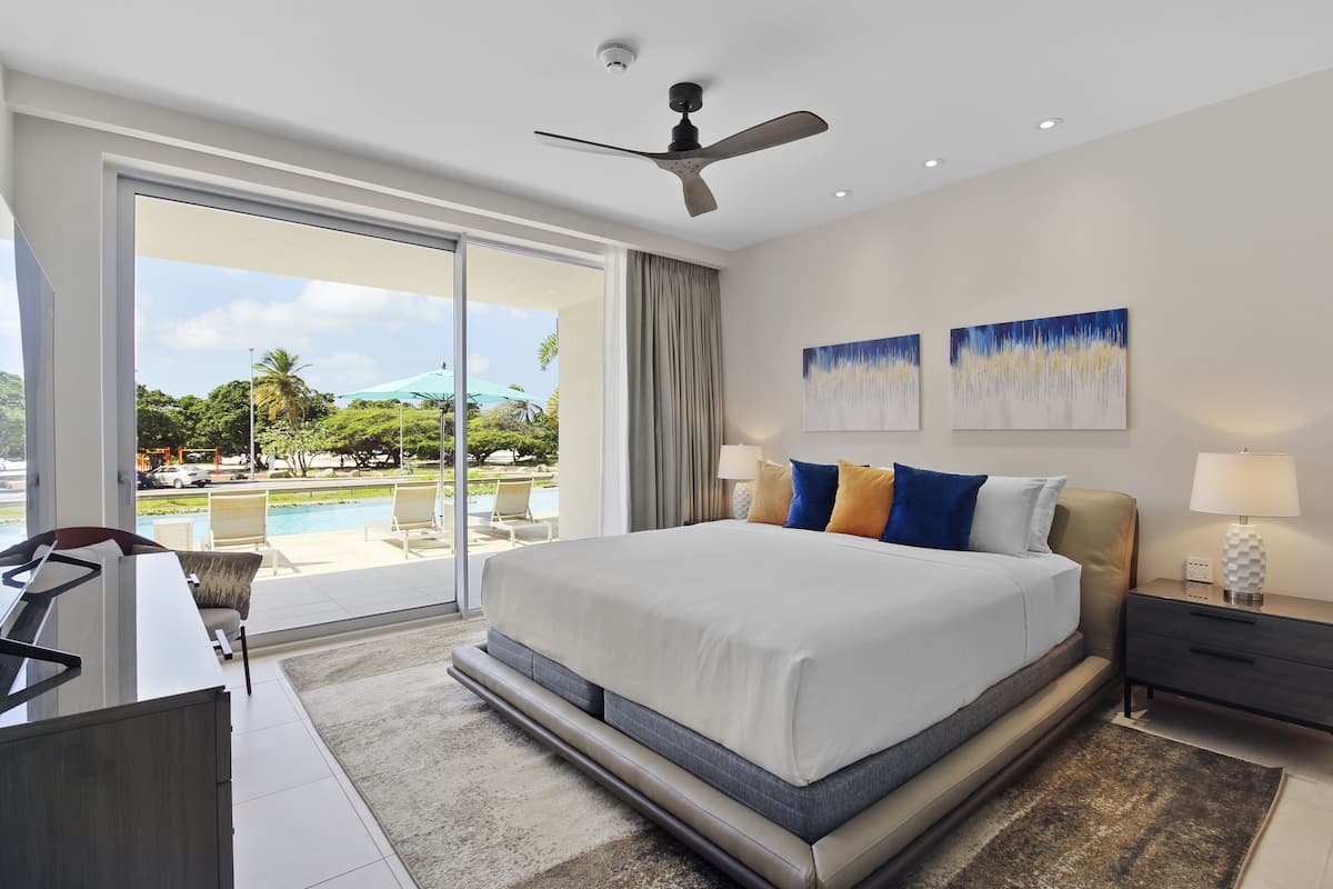 A modern hotel room with a large bed, a desk, and a view of a pool and greenery.