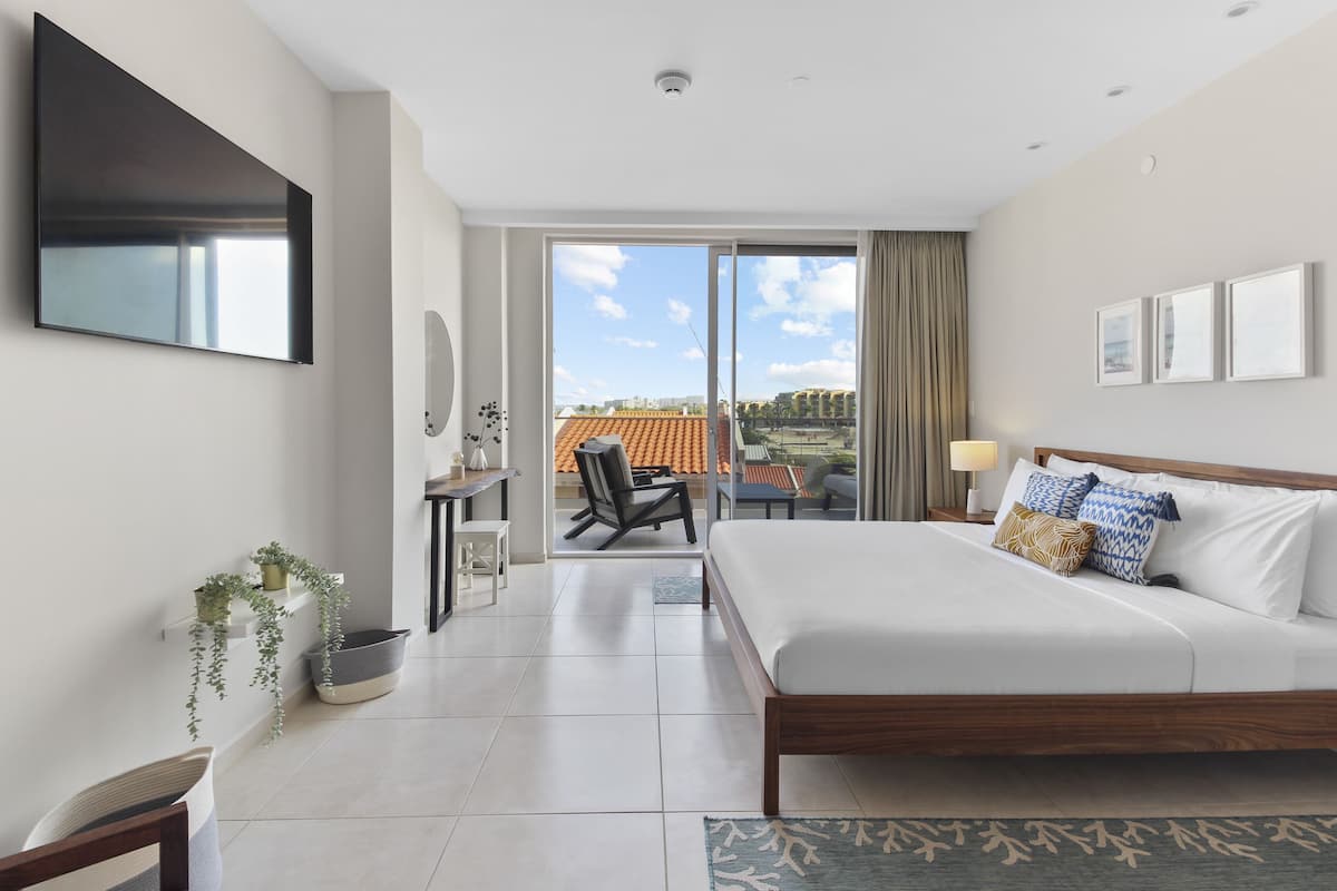 A modern hotel room with a large bed, a TV mounted on the wall, a desk with a chair, and a view of the outside.