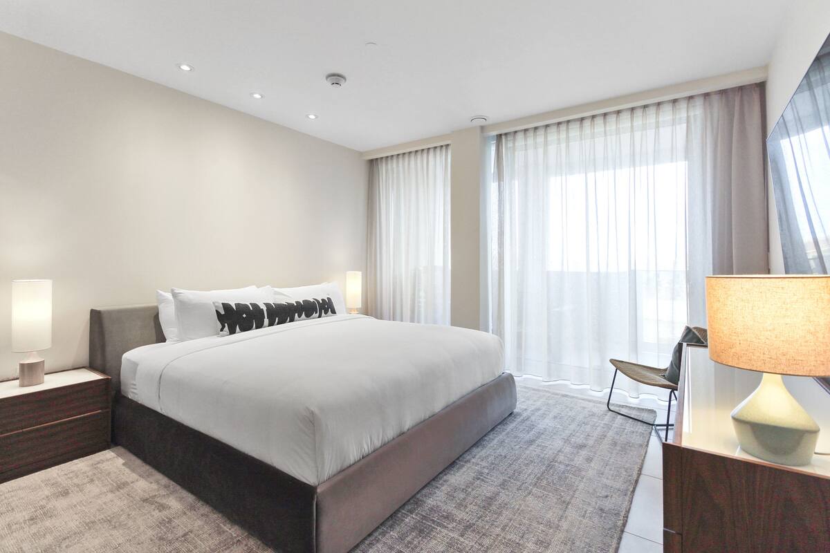 A modern hotel room with a large bed, a nightstand, a desk with a lamp, and a view of the outdoors through sheer curtains.