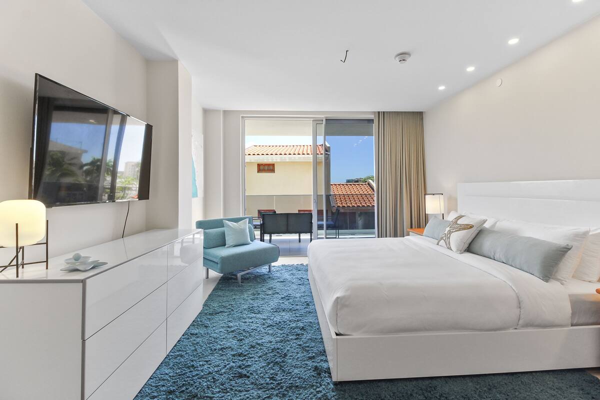 A modern hotel room with a large bed, a TV, a desk, and a balcony with a view.