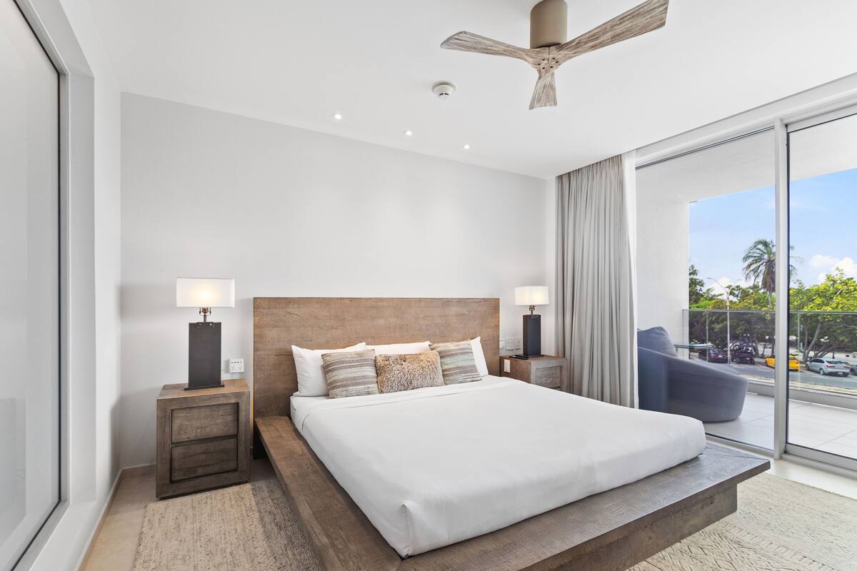 A modern hotel room with a large bed, wooden headboard, bedside lamps, and a view of the outdoors through a large window.