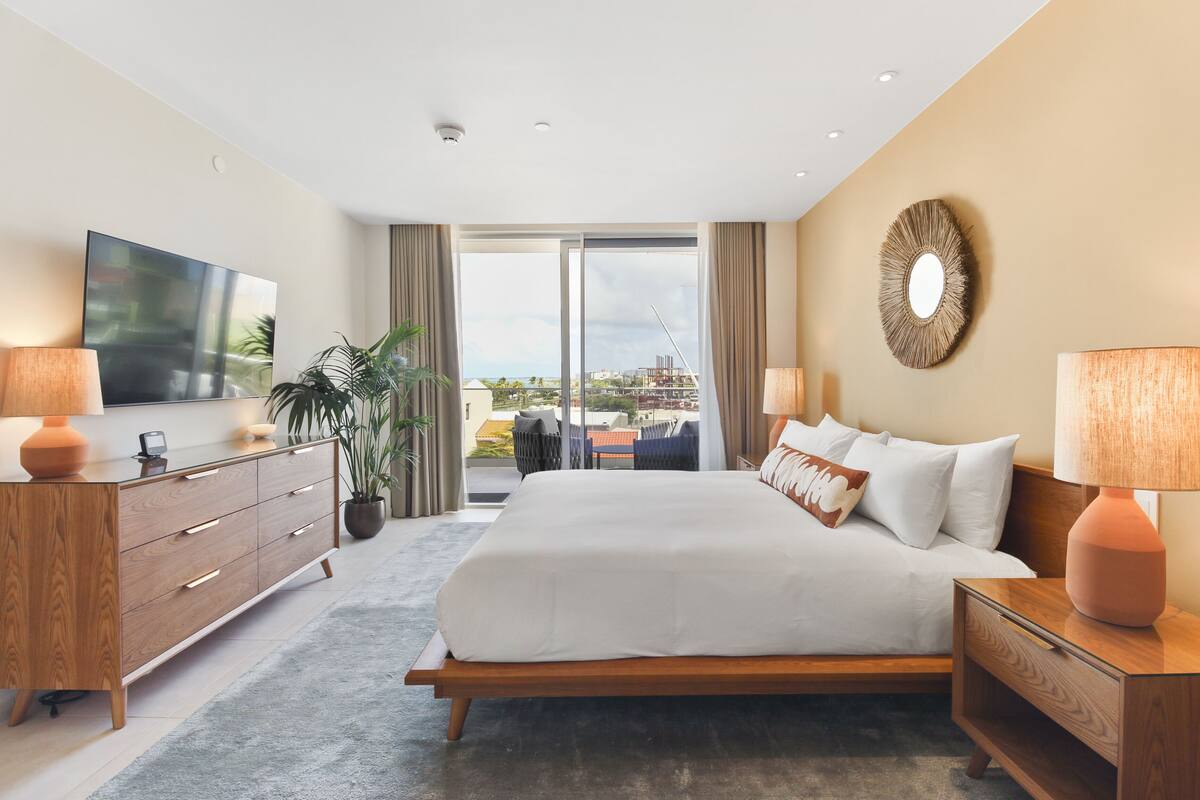 A modern hotel room with a large bed, a TV, a wooden dresser, and a balcony with a view.