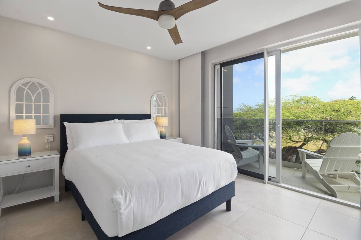 A bedroom with a large bed, two bedside lamps, a ceiling fan, and a balcony with chairs.