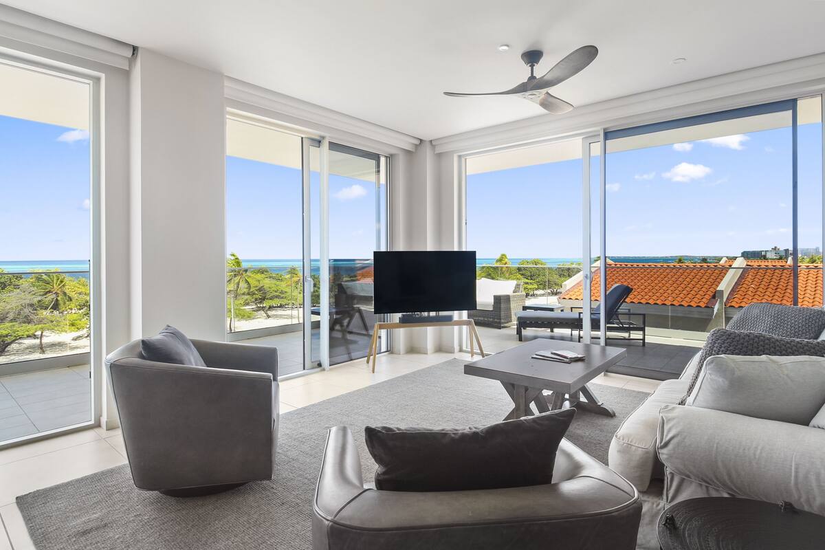 A modern living room with a flat-screen TV, grey sofas, and a large window overlooking a coastal view.