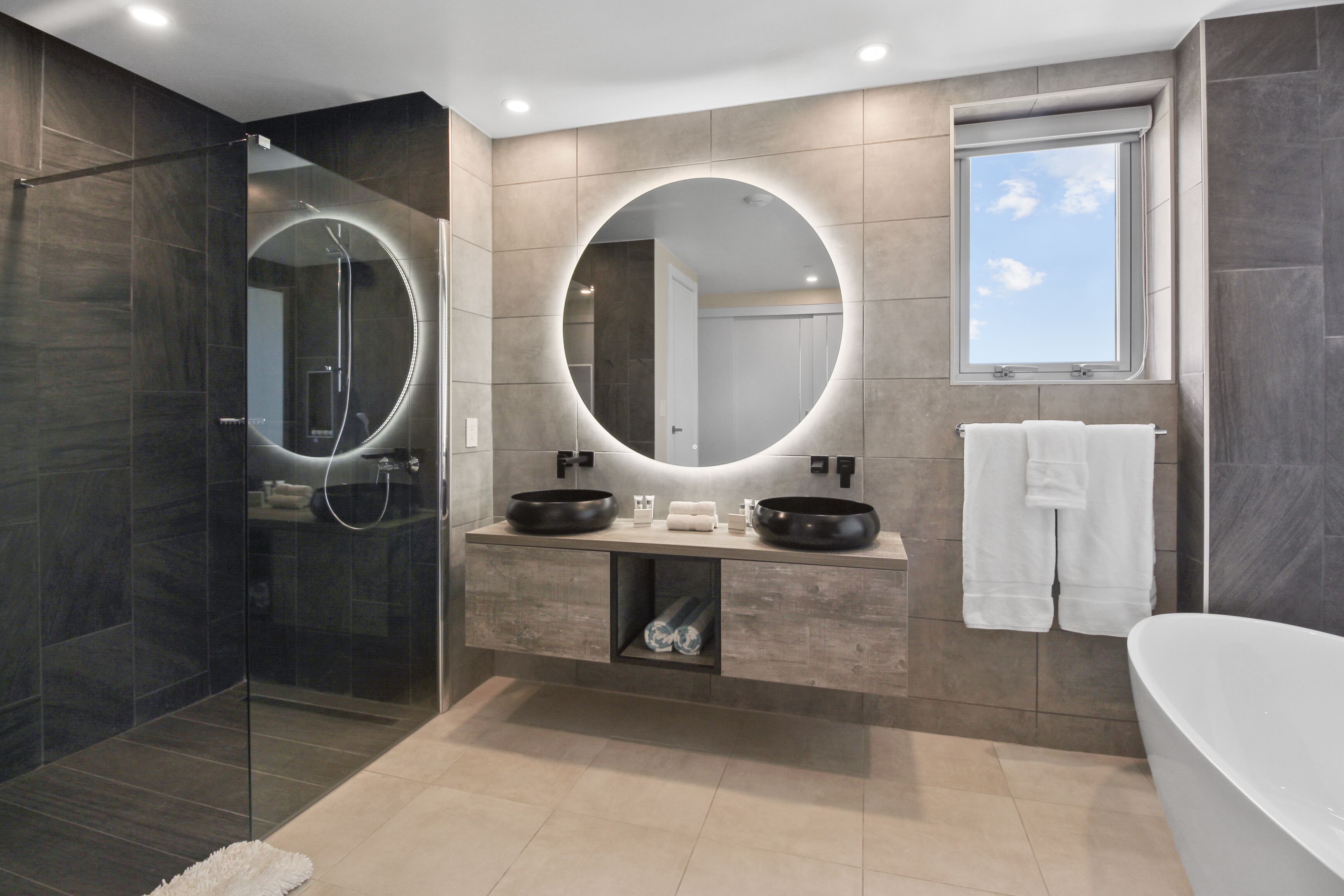 A modern bathroom with a glass-enclosed shower, two sinks with round mirrors, a bathtub, and a window with a view of the sky.