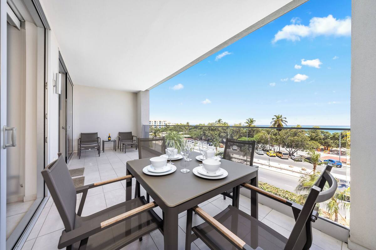 A balcony with a table set for four, chairs, and a view of the street and palm trees.