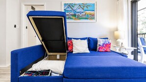 A room with a blue sofa bed, a painting on the wall, a lamp, and a chair.