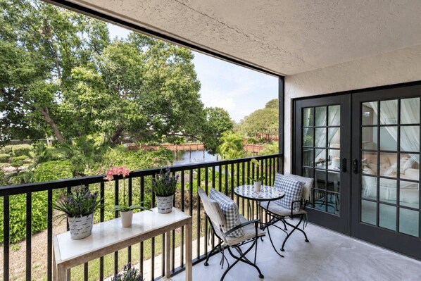 A balcony with a table and chairs, potted plants, and a view of a garden and water body.