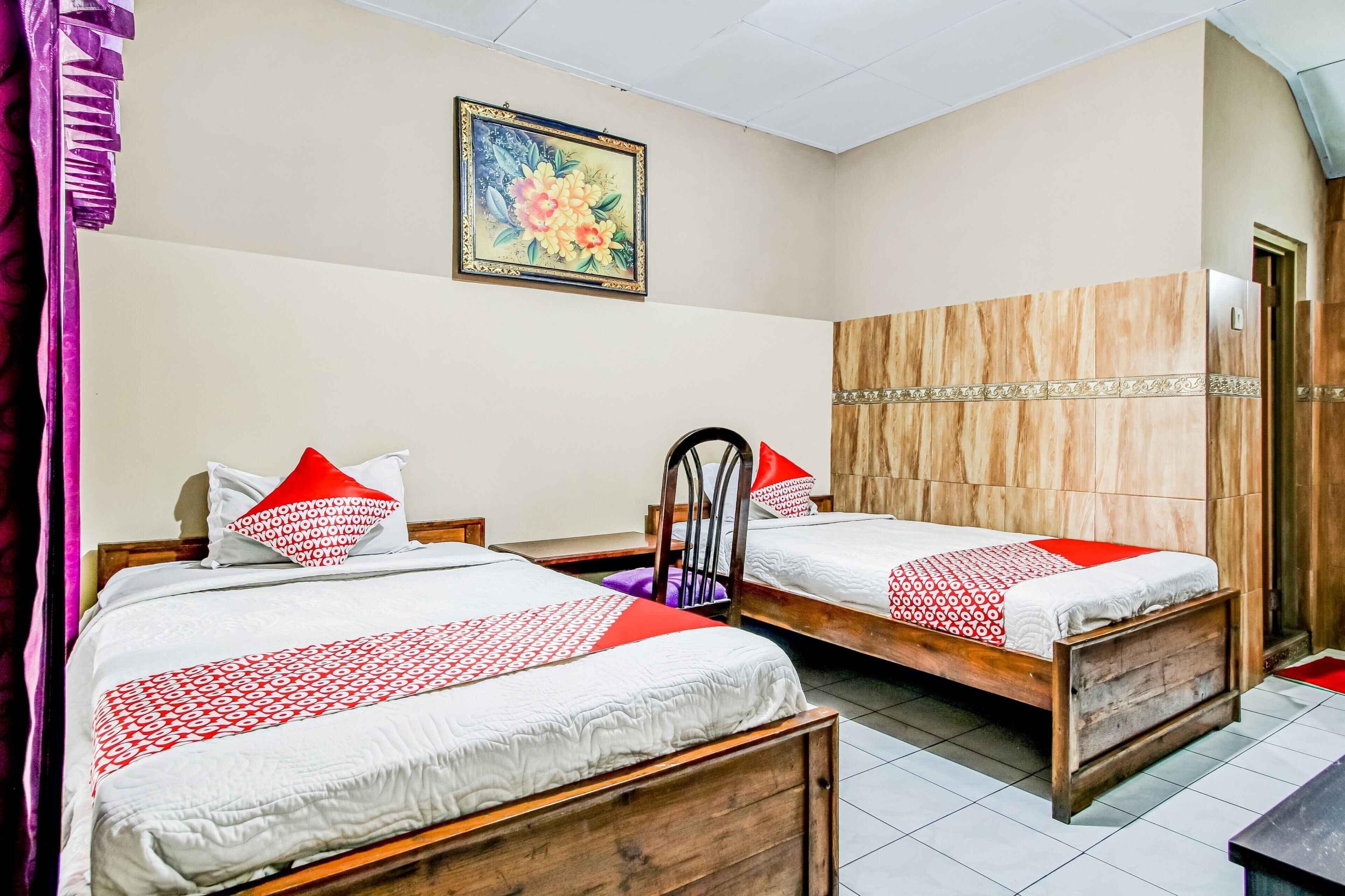 A hotel room with two wooden beds, a wooden wardrobe, a framed floral artwork on the wall, and a purple curtain.