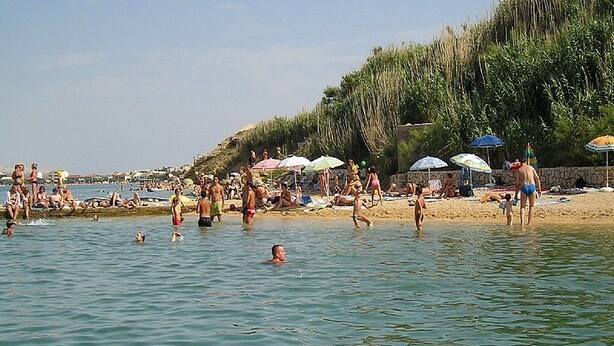 Una playa con gente nadando y tomando el sol, sombrillas y un muro de piedra.