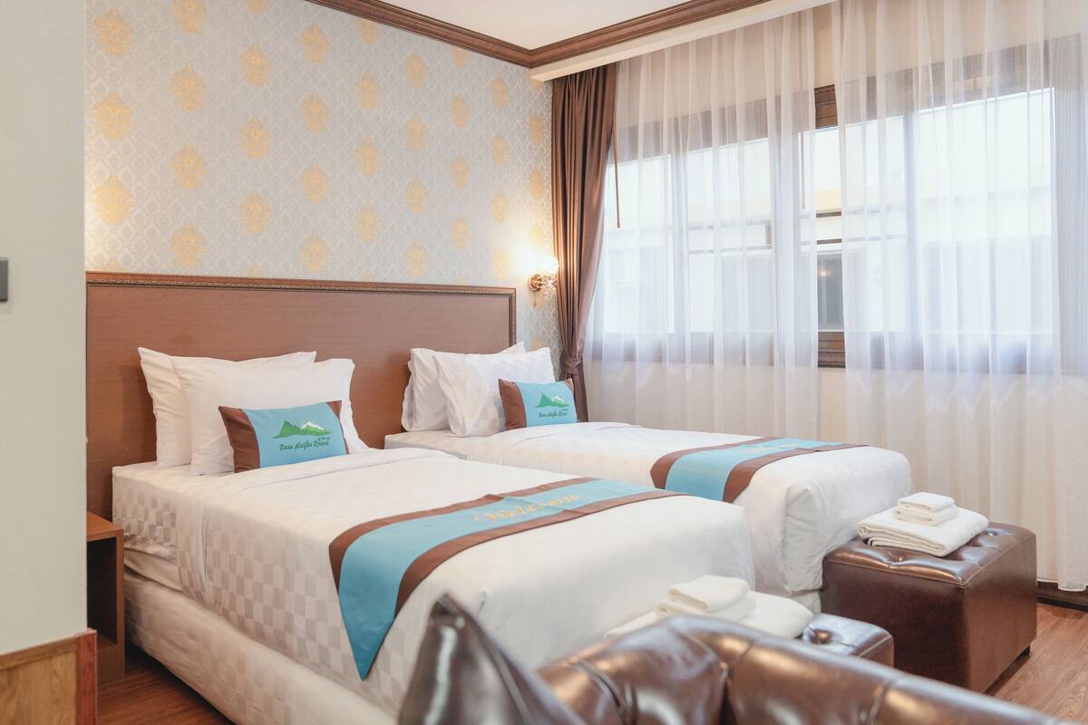 A hotel room with two beds, a wooden headboard, a large window with sheer curtains, and a checkered bedspread.