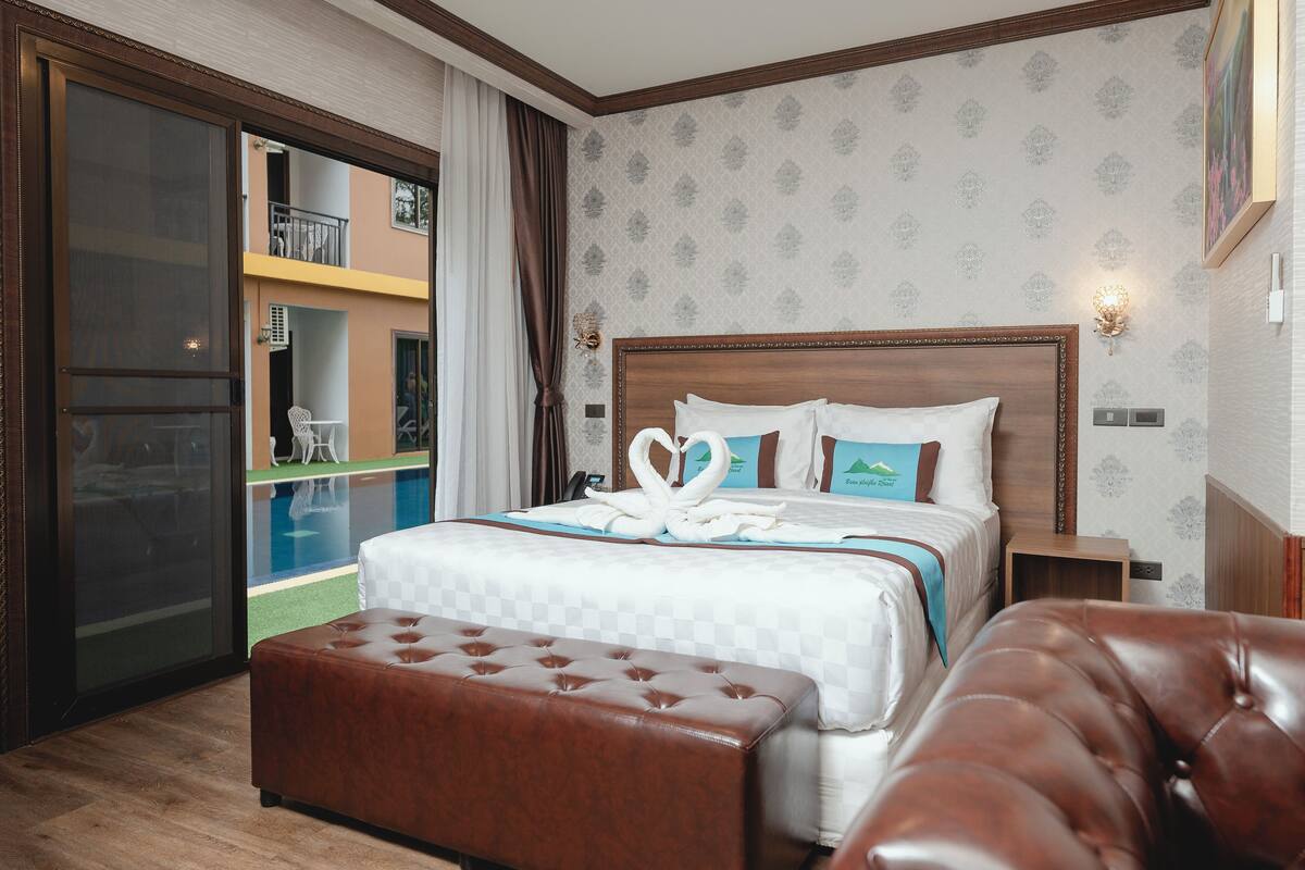 A hotel room with a large bed, a wooden headboard, a brown leather bench, and a balcony with a view of the pool.