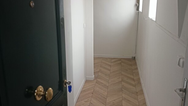 Ein Flur mit Holzboden im Fischgrätmuster, weißen Wänden, einer dunklen Tür mit Messingklinke und einem Fenster mit weißer Jalousie.