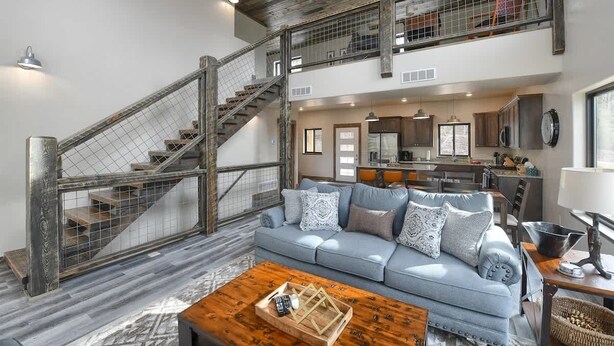 Ein großzügiger Wohnbereich mit hoher Decke, einer Treppe mit Metallgeländer, einem hellblauen Sofa mit gemusterten Kissen und einem Holztisch.