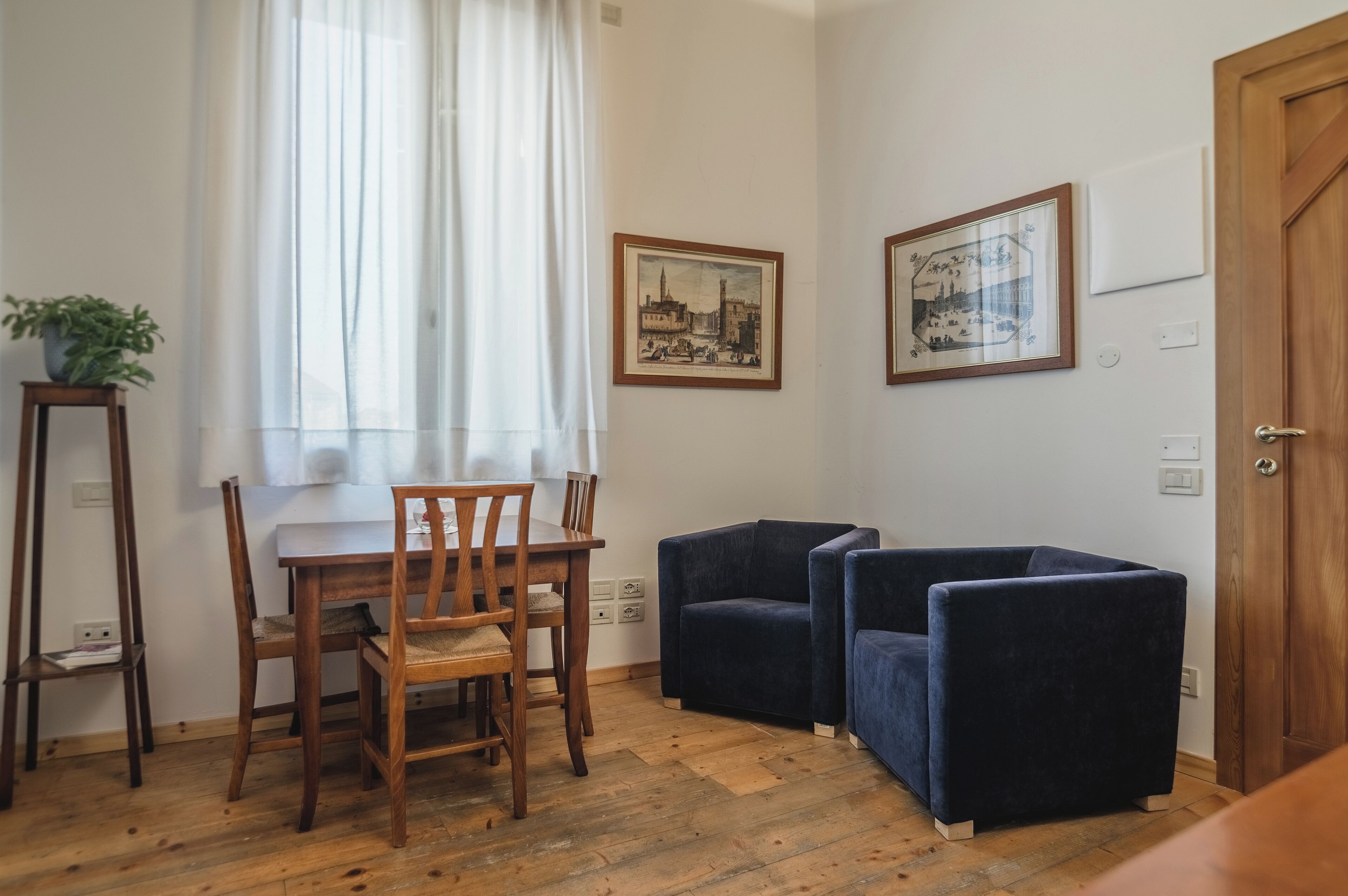 Una habitación con una mesa de comedor de madera y sillas, dos sillones azul oscuro, una planta en maceta y dos cuadros en la pared.