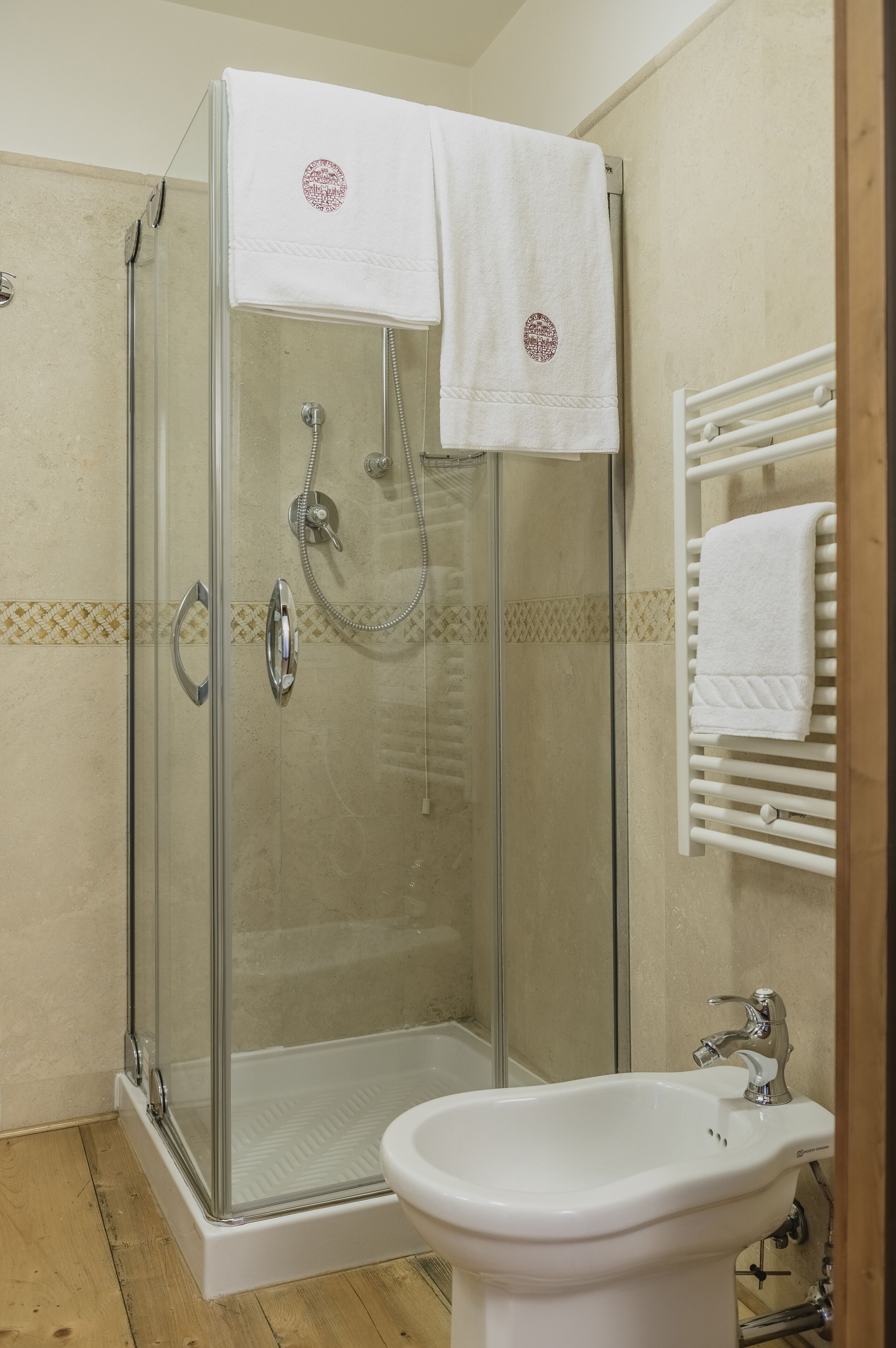 Ducha envuelta en vidrio, con cabezal de ducha manual y grifo empotrado en la pared. Hay un lavabo blanco con grifo cromado y un espejo encima.