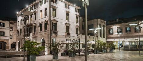 A spacious square with a checkered floor, surrounded by buildings with classical architecture and outdoor seating areas.