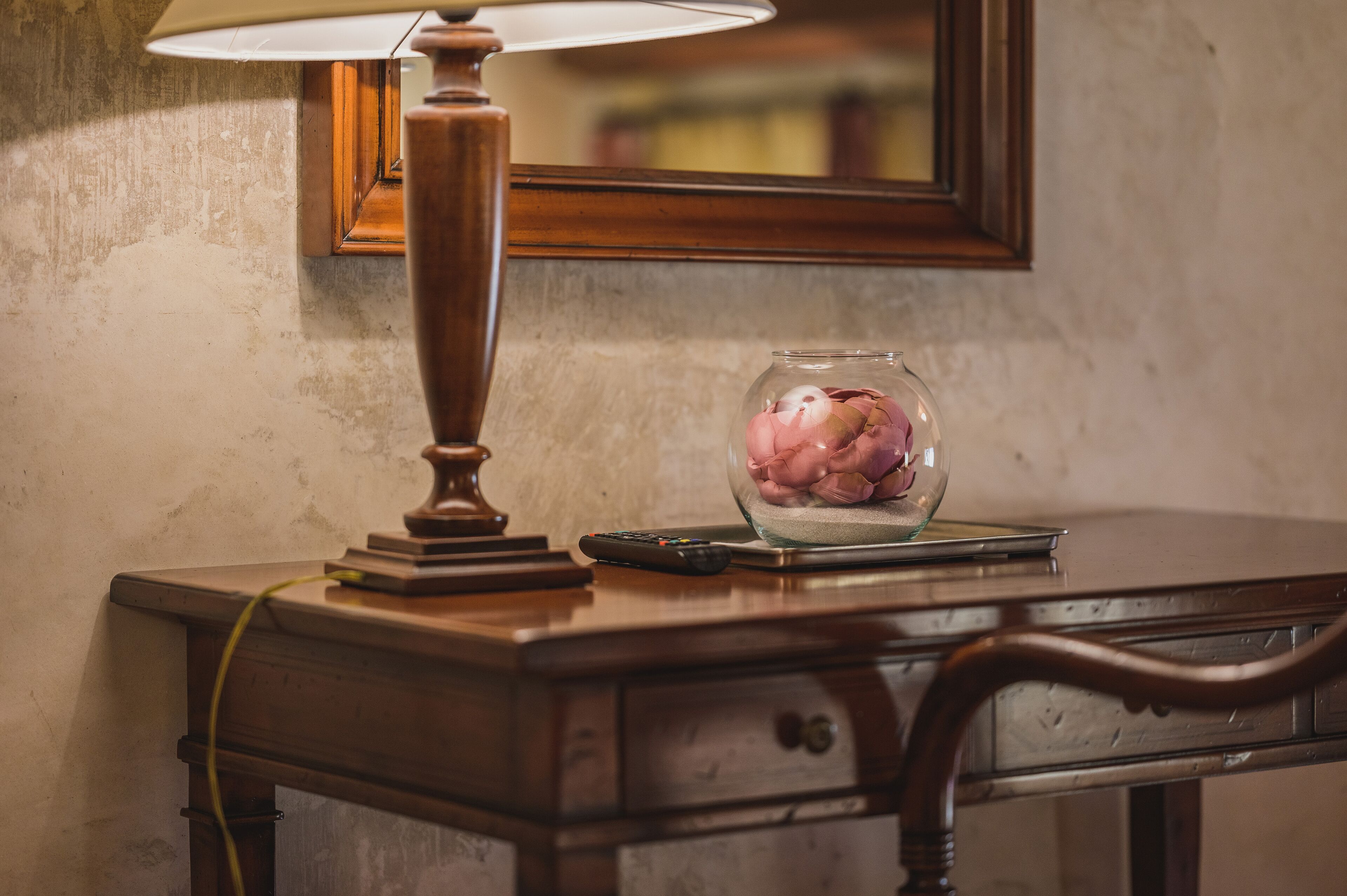 A wooden desk with a lamp, a glass vase with flowers, and a remote control.