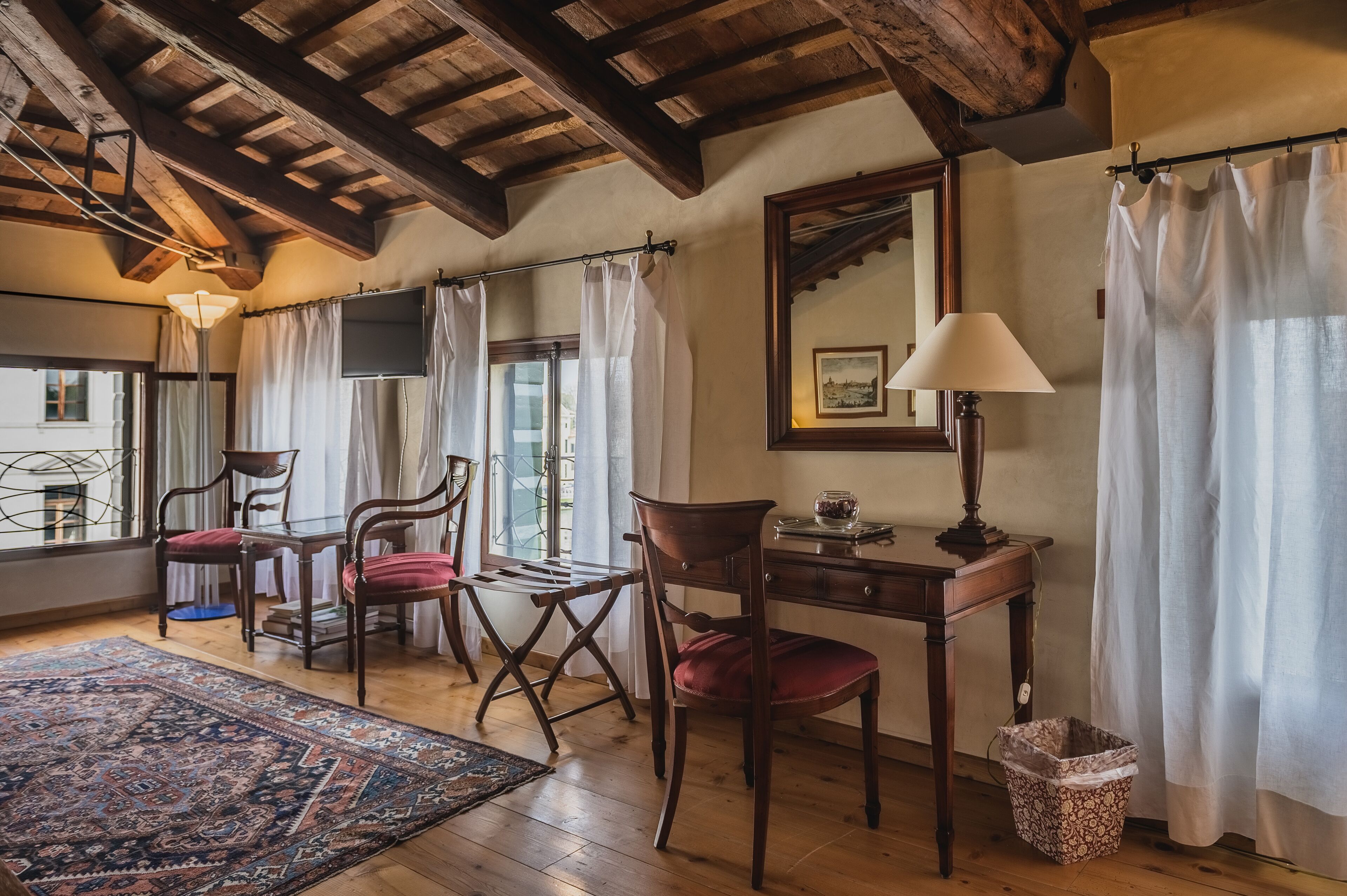 A room with wooden beams, a desk with a lamp, two chairs, a rug, and a window with curtains.