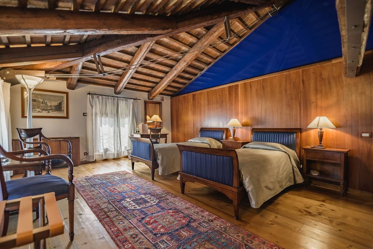 Een slaapkamer met houten balkenplafond, twee bedden, een bureau, stoelen en een kamerkleed met patroon.