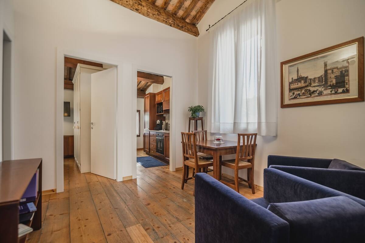 Een ruime woonkamer met houten vloer, een eethoek met tafel en stoelen, en een keuken die zichtbaar is door een open doorgang.
