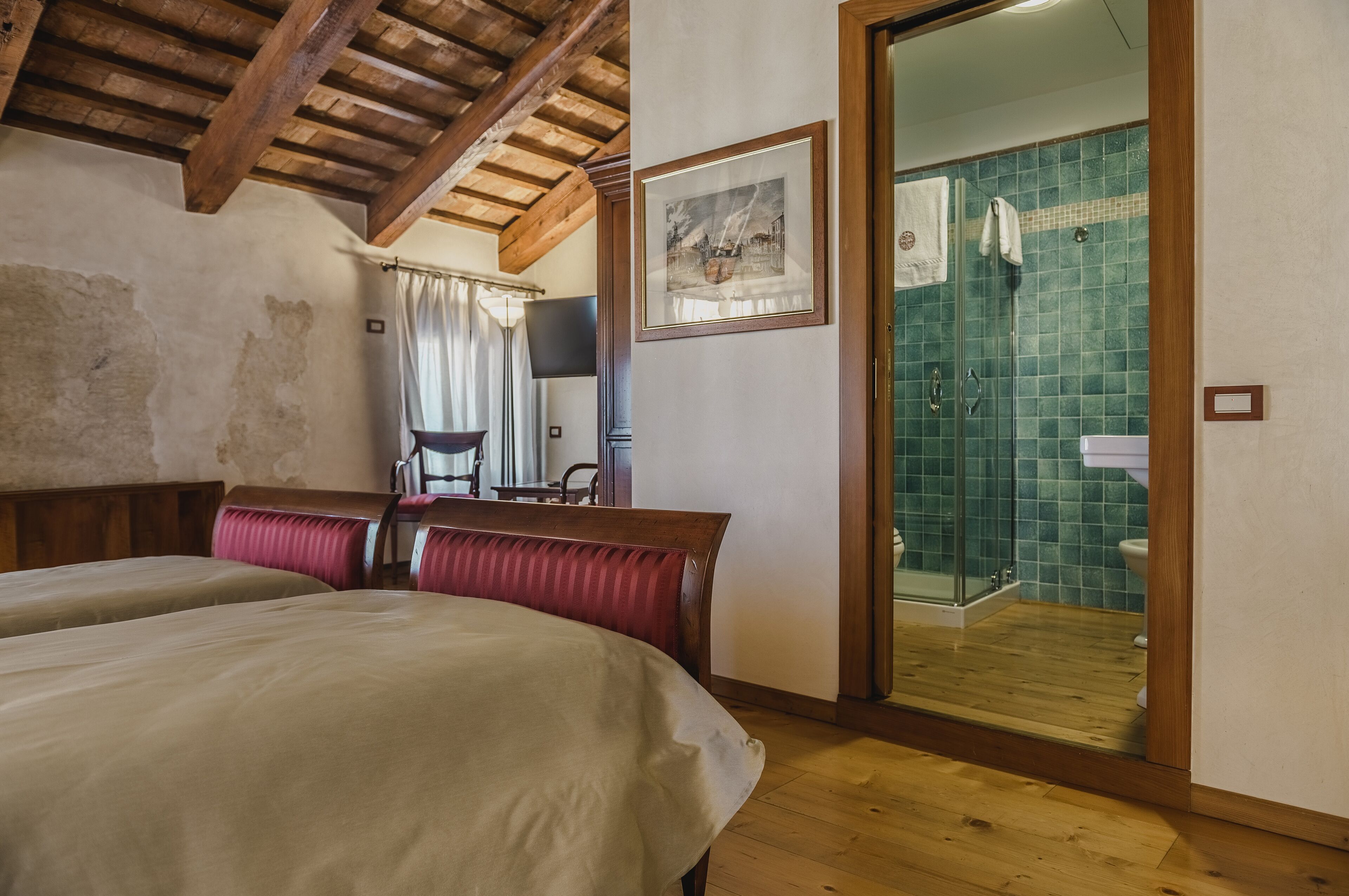 A bedroom with two beds, a wooden ceiling, a painting, and a bathroom with a green tiled wall and a shower.