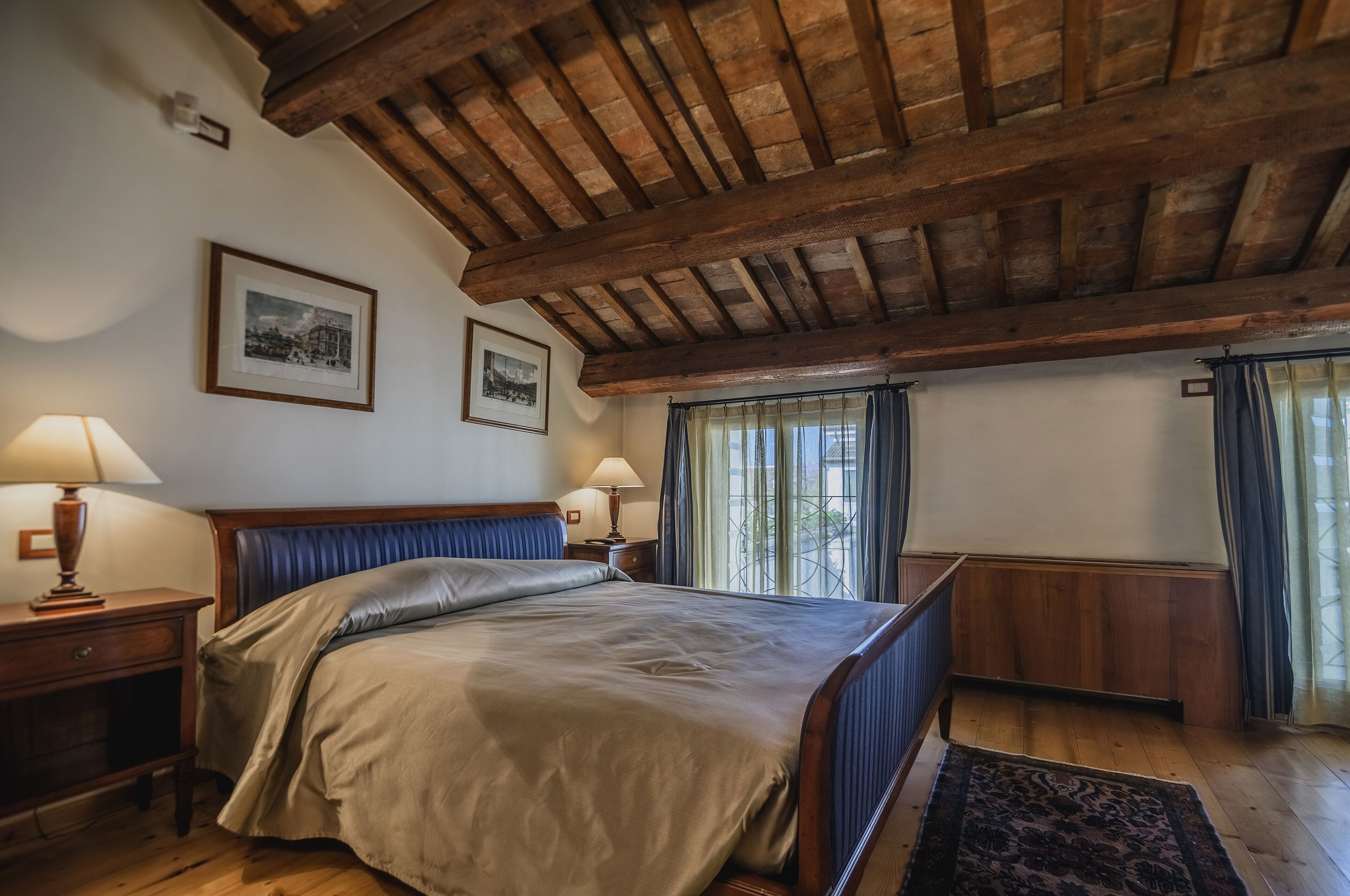 A bedroom with a wooden ceiling, a bed with a beige bedspread, two bedside lamps, a nightstand, a framed picture on the wall, and a window with curtains.
