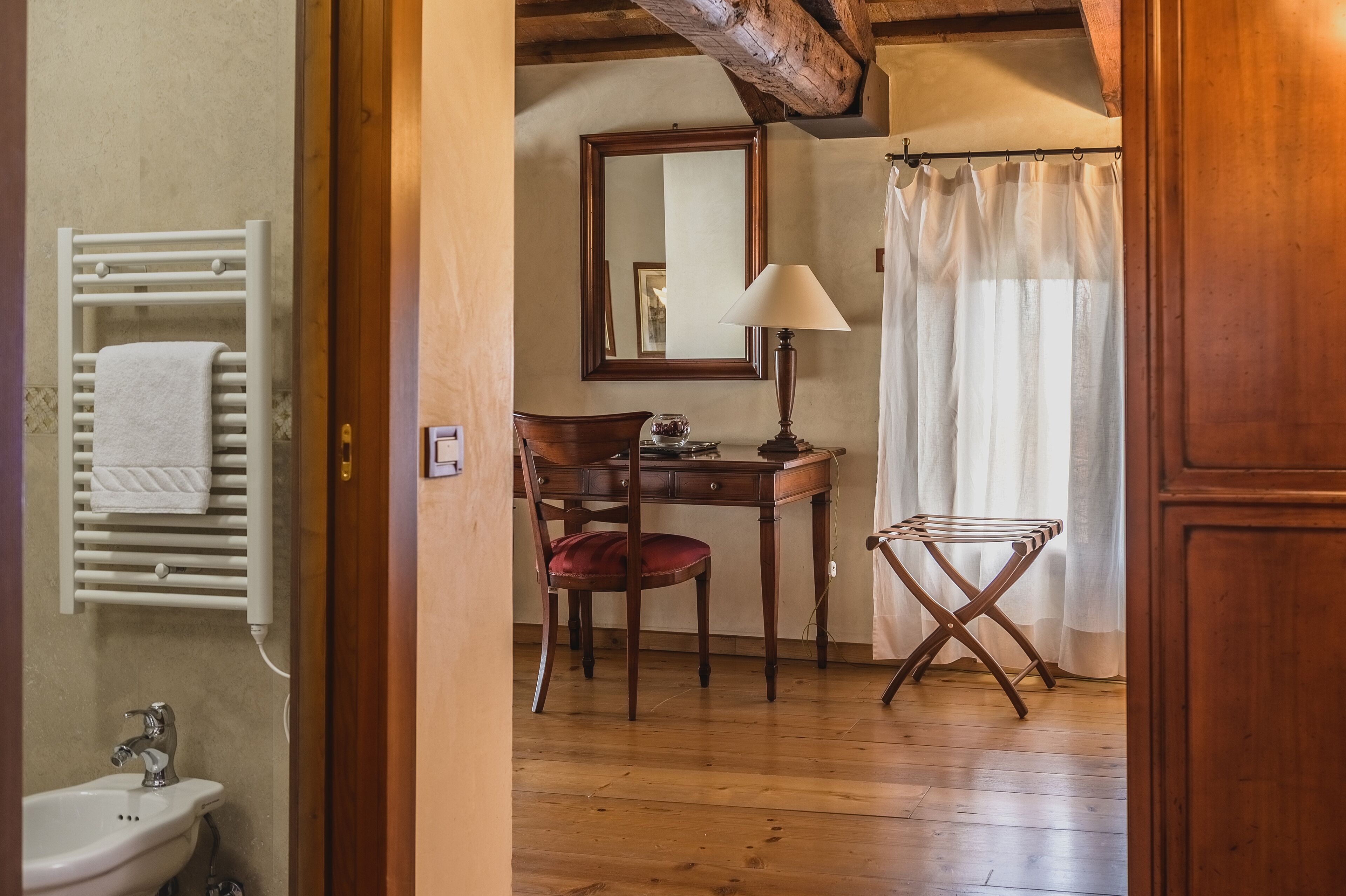 A wooden floor, a wooden chair, a desk with a lamp, a mirror, a towel rack, and a shower curtain.
