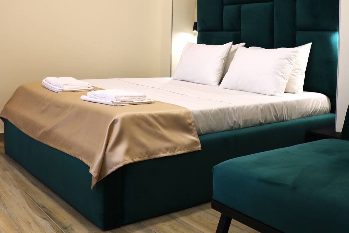 Una cama bien tendida con sábanas blancas y un plaid color beige, un cabecero tapizado en verde y un banco a juego al lado de la cama.