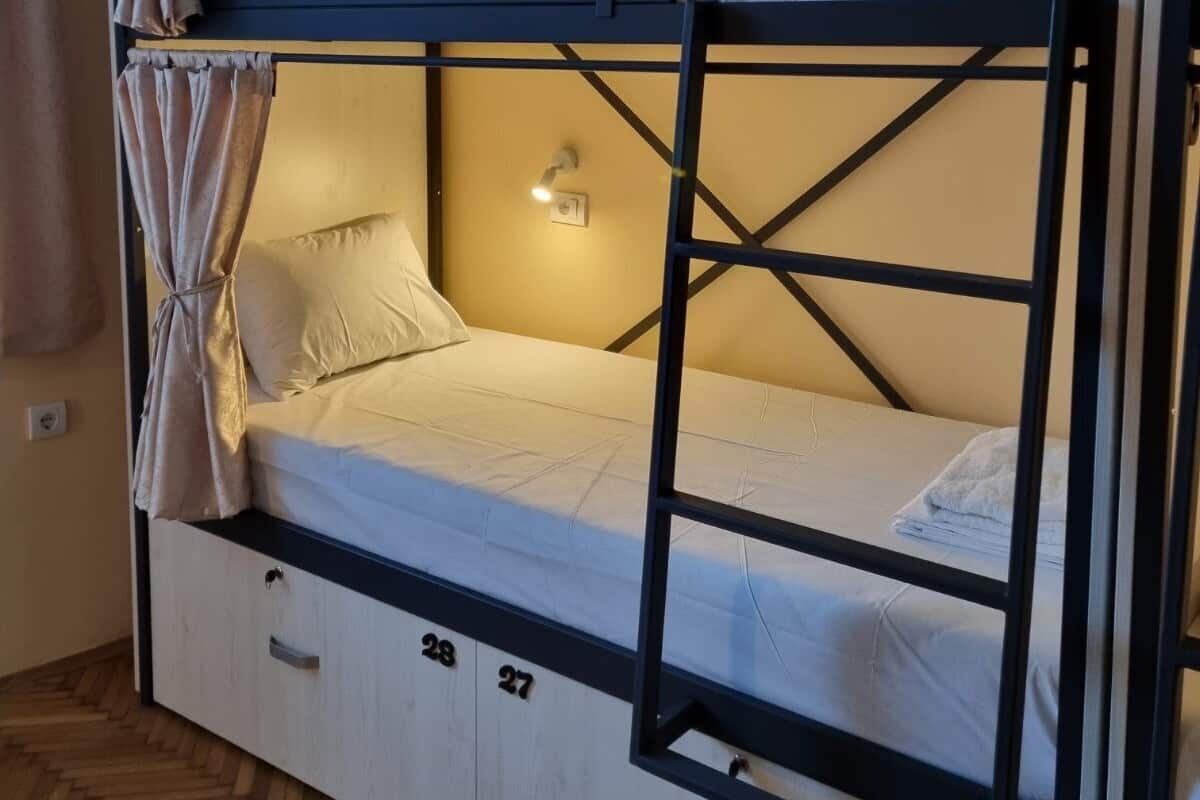 Una cama litera con dos niveles, cada uno con almohadas y una colcha. Hay un estante pequeño con una luz, y una cortina atada al lateral de la cama.