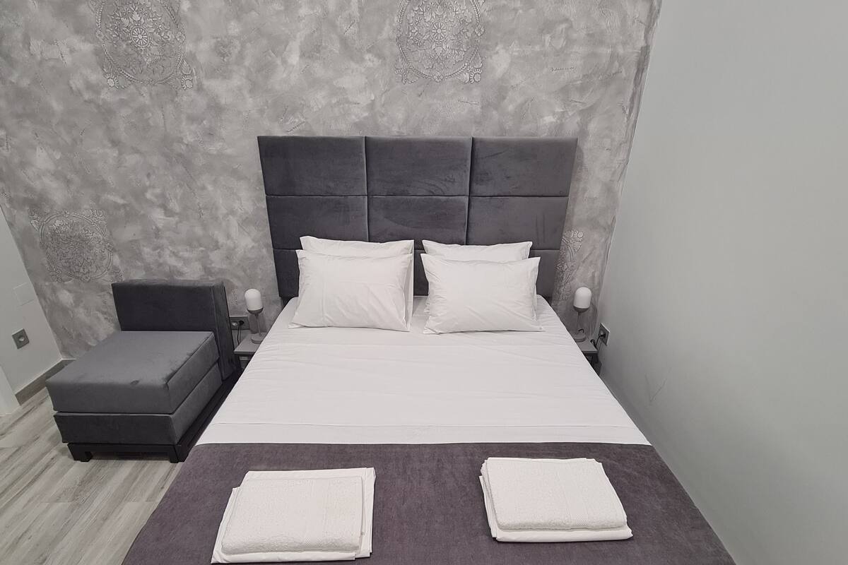 Una cama bien hecha con dos almohadas, una mesita de noche con una lámpara y una silla pequeña.