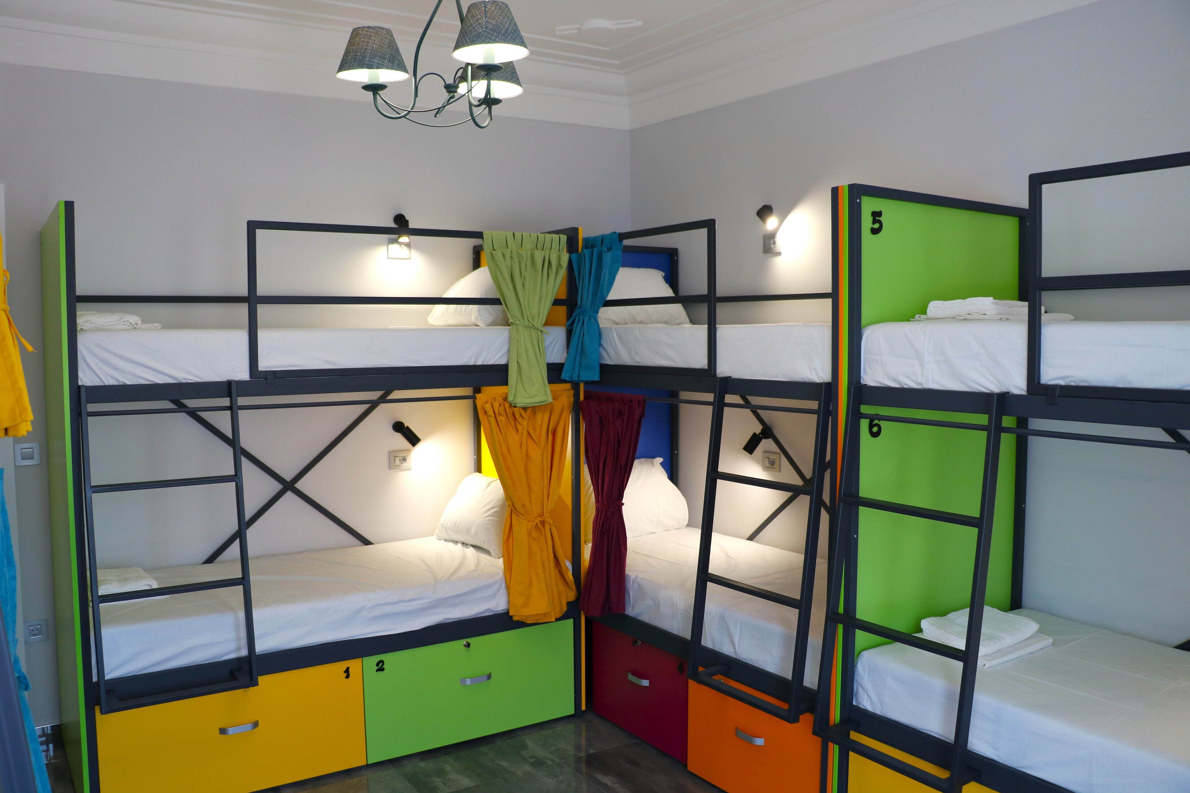 Habitación con literas, tres en total, cada una con un color diferente en la cama inferior. Hay luces fijadas en la pared sobre las camas y una lámpara colgante.