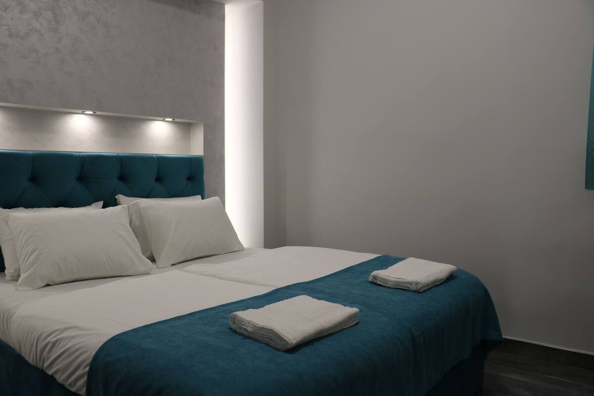 Una habitación de hotel con una cama grande, dos almohadas, un cabecero verde azulado y una pared blanca.