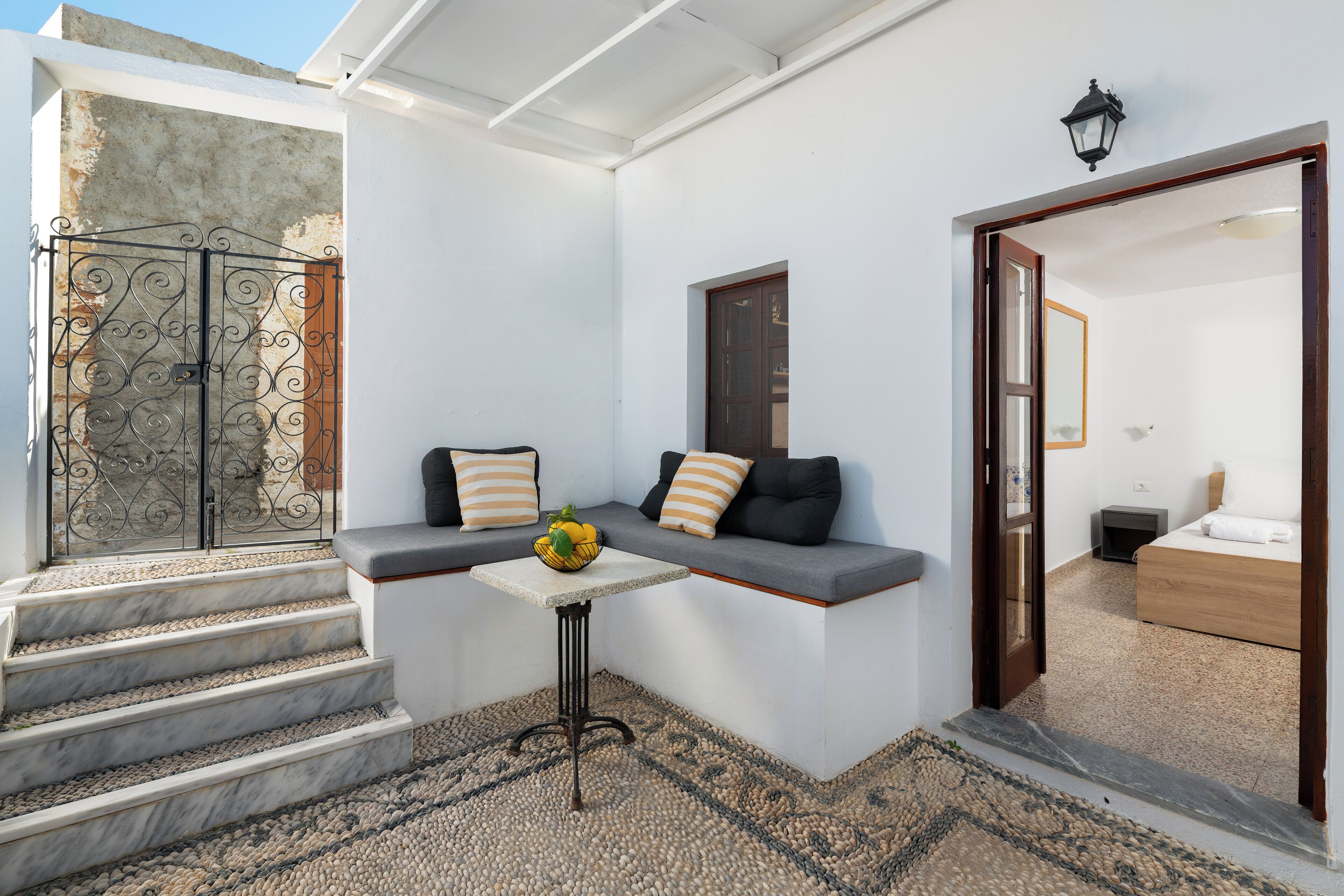 Un intérieur moderne avec un escalier en pierre, un coin salon avec des coussins, une petite table sur laquelle est posé un panier de fruits, et une chambre visible à travers une porte ouverte.