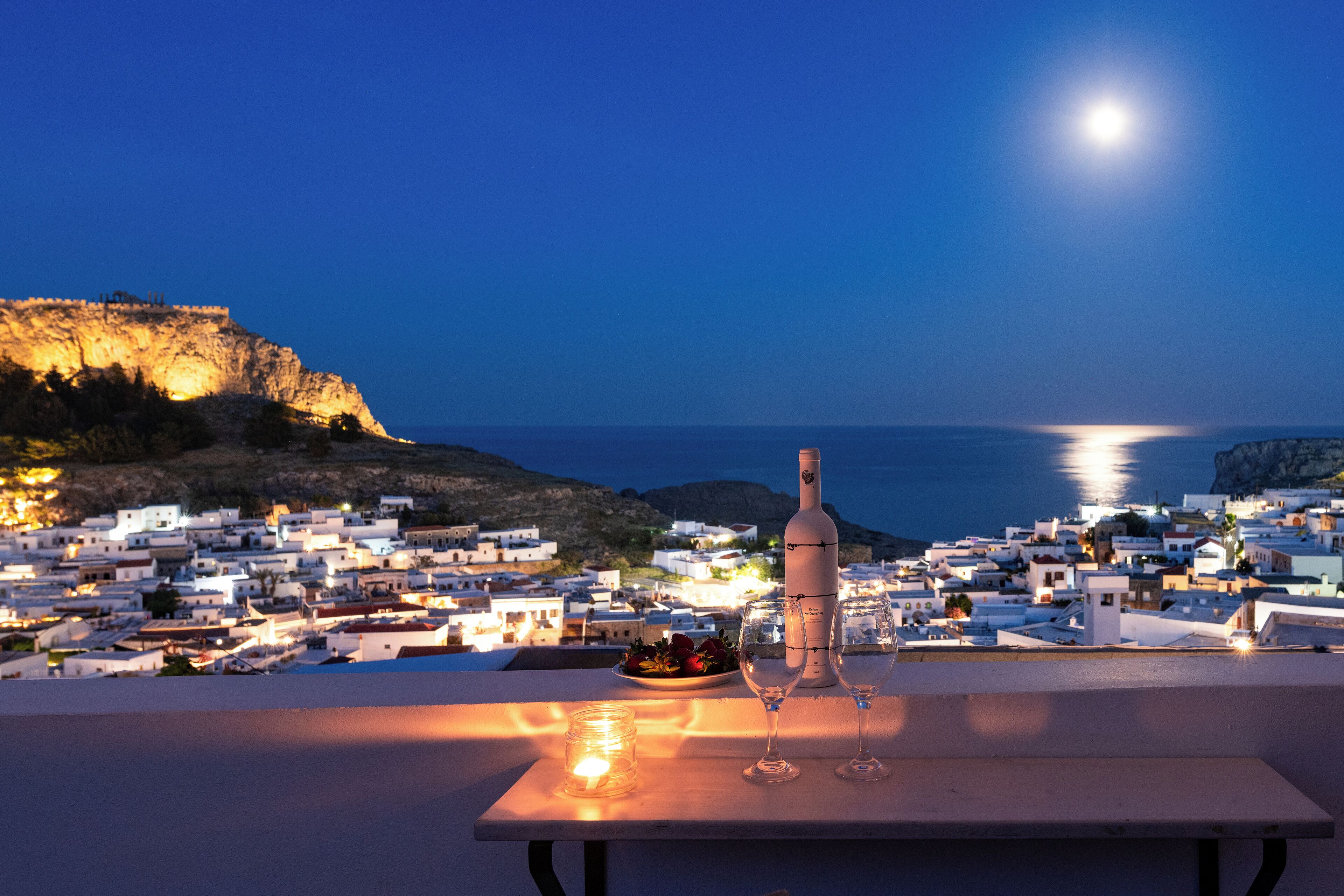A table with a bottle and glasses, overlooking a coastal town at night.
