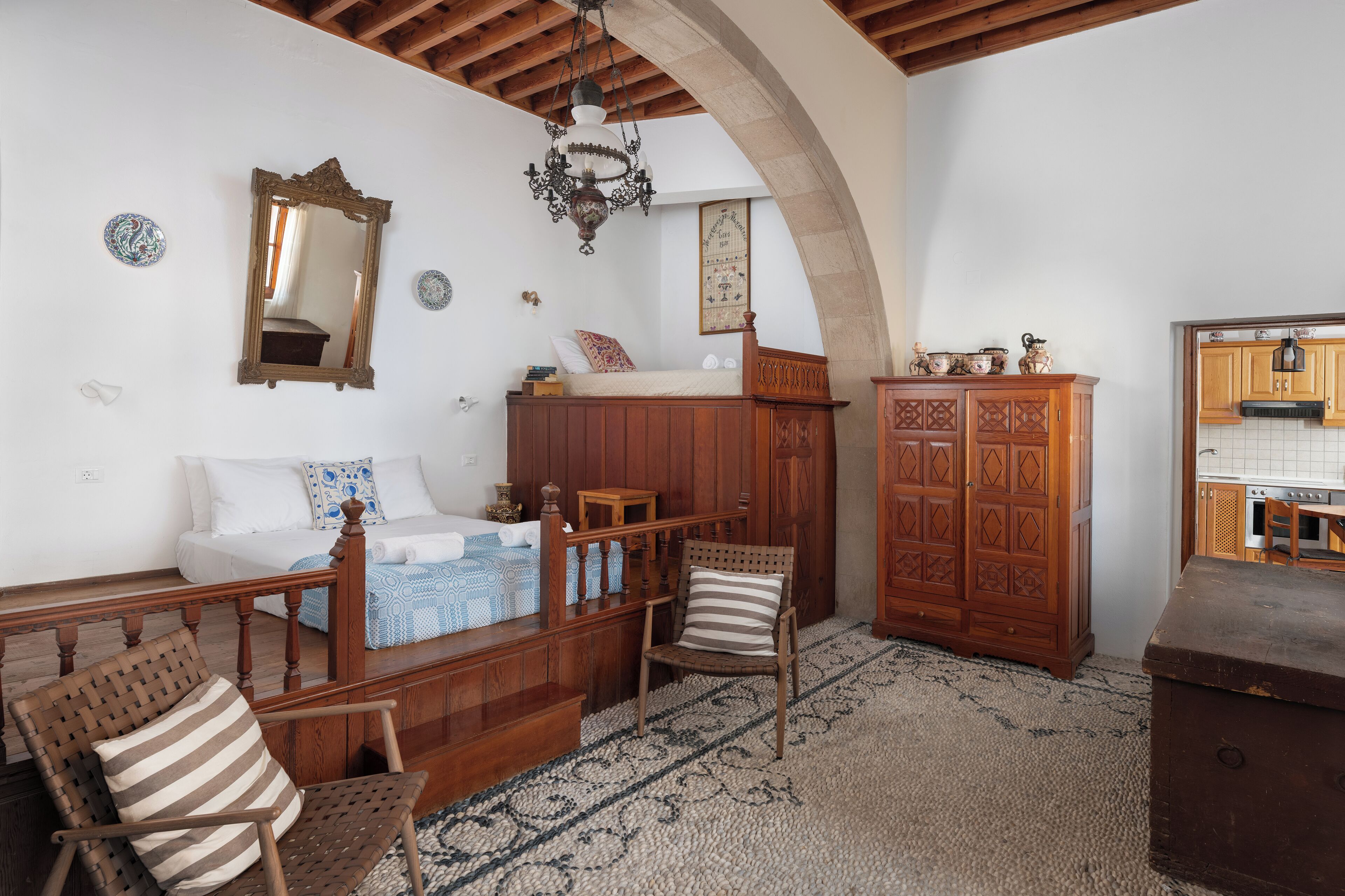 A bedroom with a wooden bed, a wooden chair, a cabinet, and a kitchen area visible through an archway.