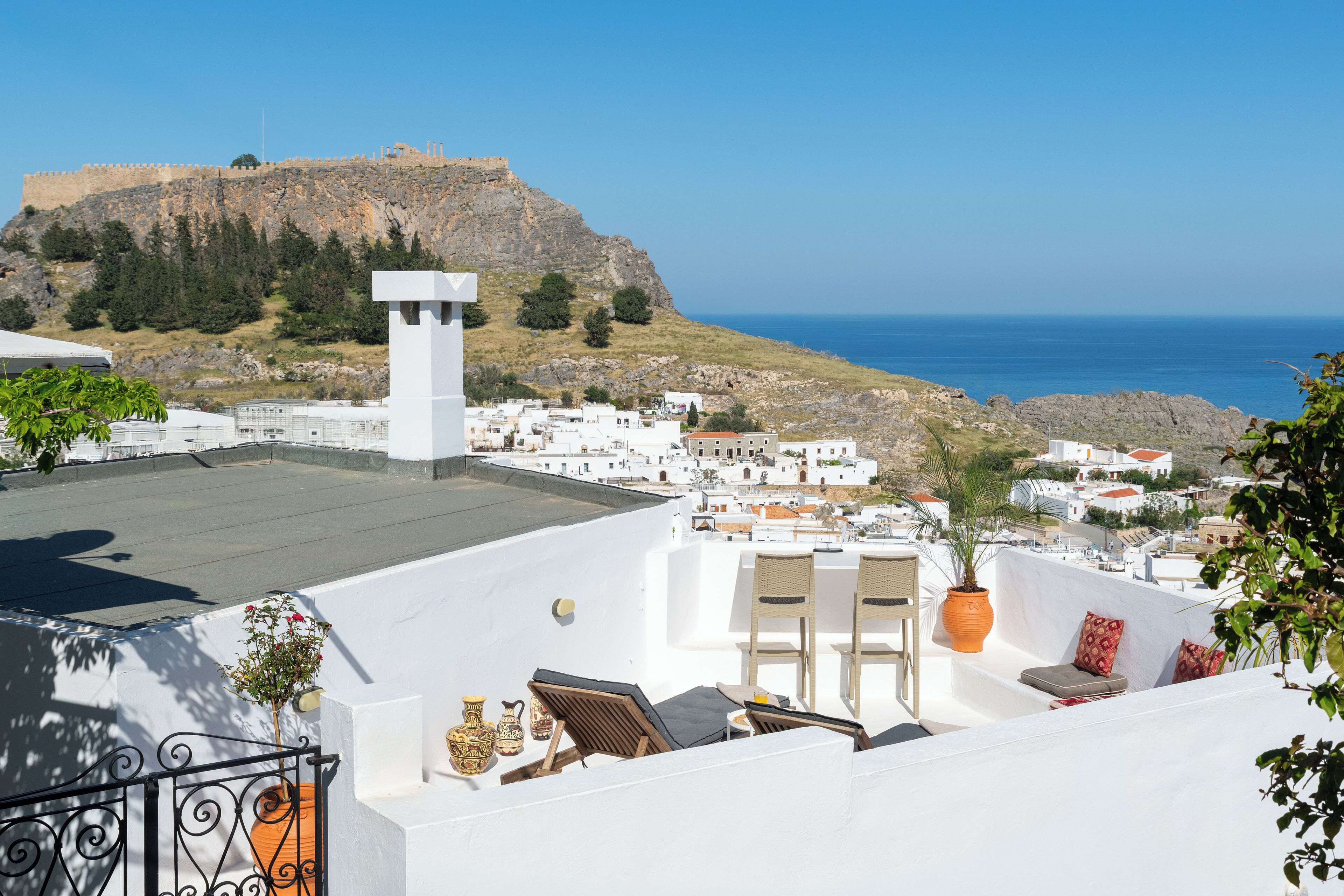 A rooftop terrace with white walls, a view of the sea, and a rocky hill in the background.