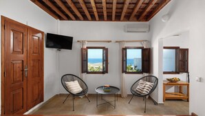 Una stanza con pavimento in legno, una TV fissata al muro, due finestre con tende, un piccolo tavolino rotondo, due sedie e un armadietto in legno.