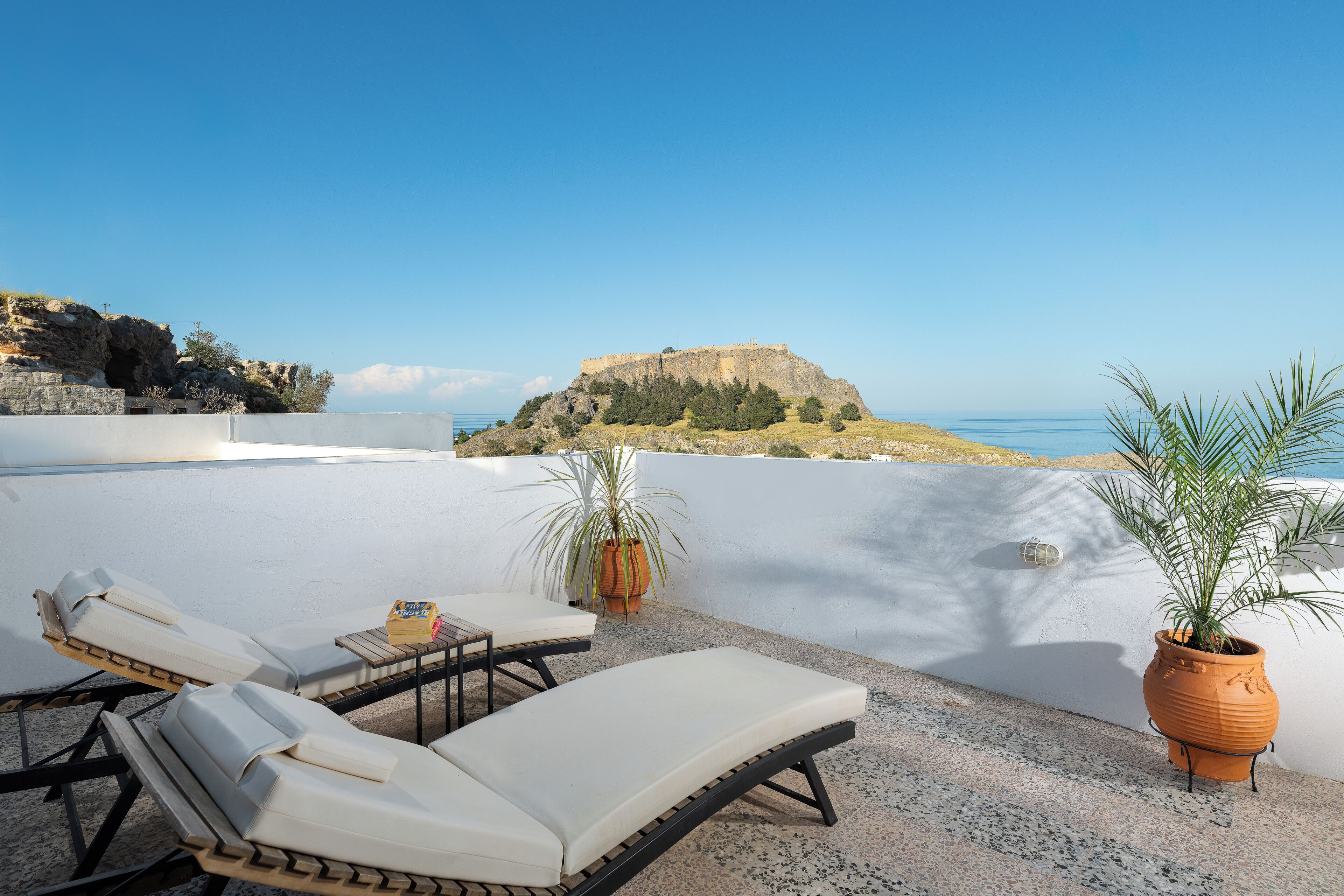Une terrasse sur le toit, aménagée avec des chaises longues, des plantes en pot et offrant une vue sur une colline rocheuse et la mer.