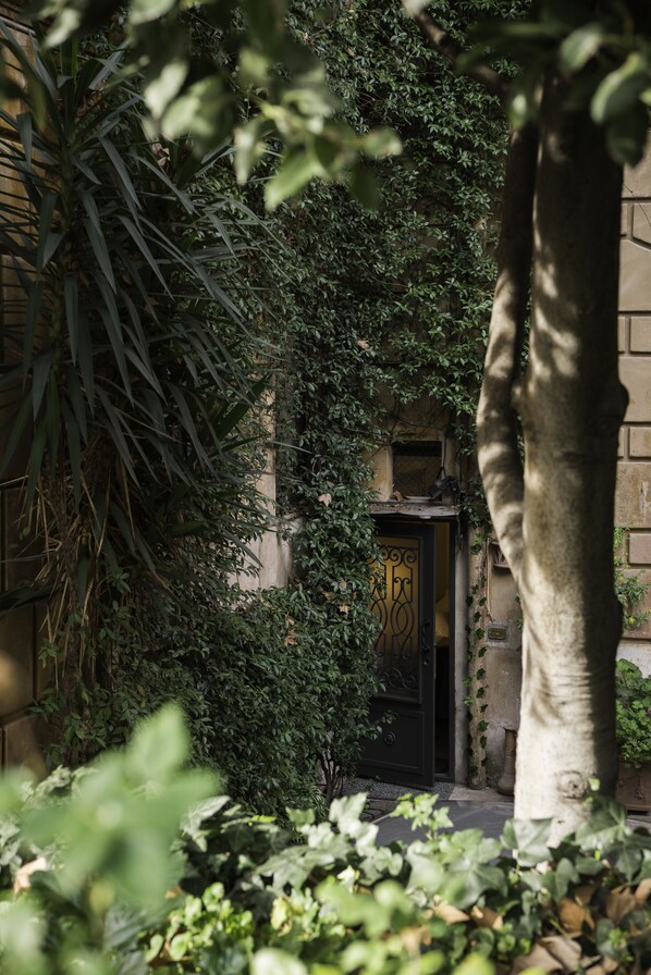 Un bâtiment doté d’une porte noire, entouré d’un feuillage dense et d’un tronc d’arbre.