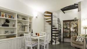 Un comedor con una mesa y sillas blancas, rodeado de muebles blancos con libros y objetos decorativos. Se ve una escalera de caracol con acabado de madera al fondo.